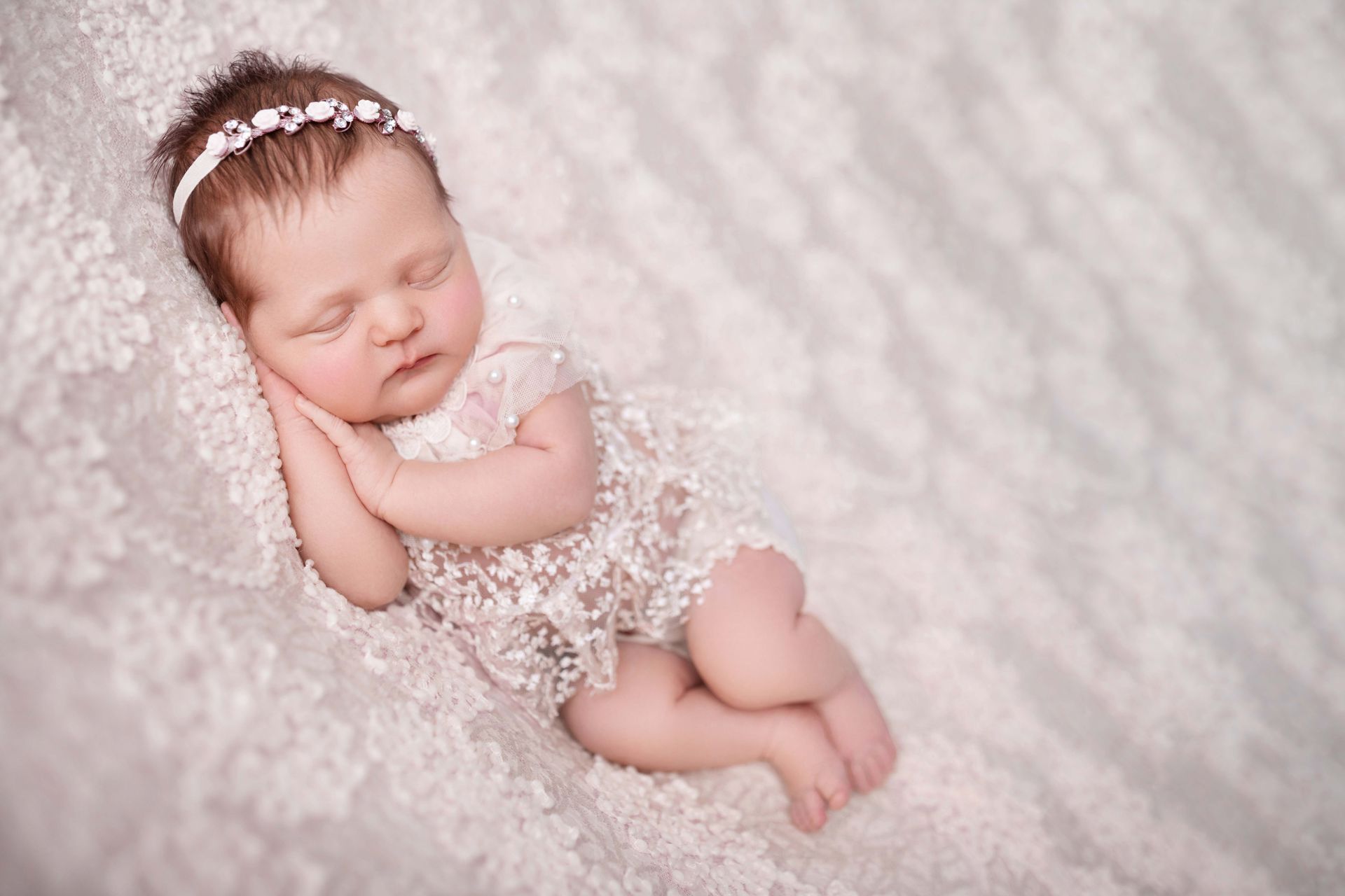 Newborn baby asleep, wearing lace outfit and floral headband, on white lacy fabric.