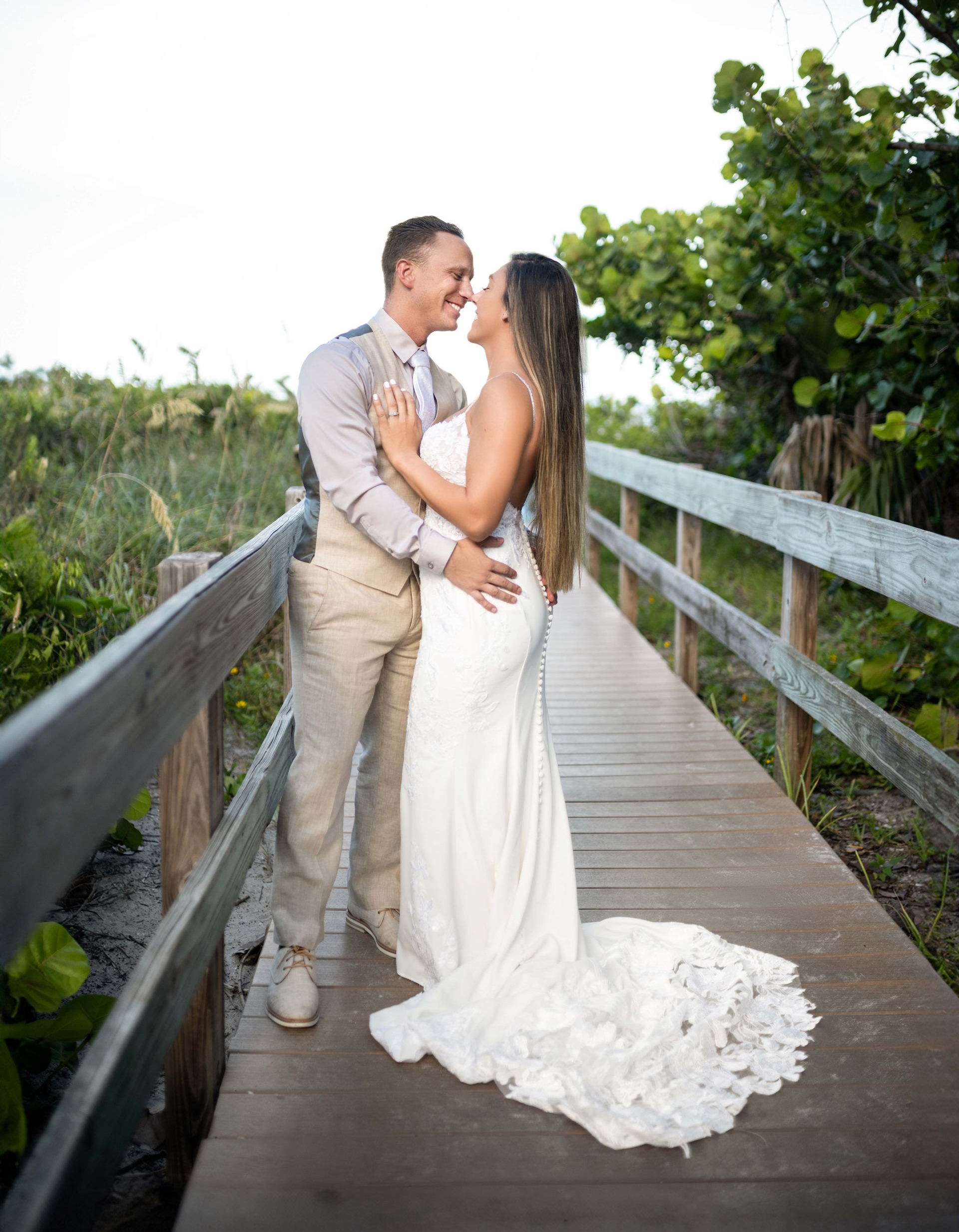 Couple embraces on a wooden bridge, bride in white gown, groom in beige suit, greenery in the background.