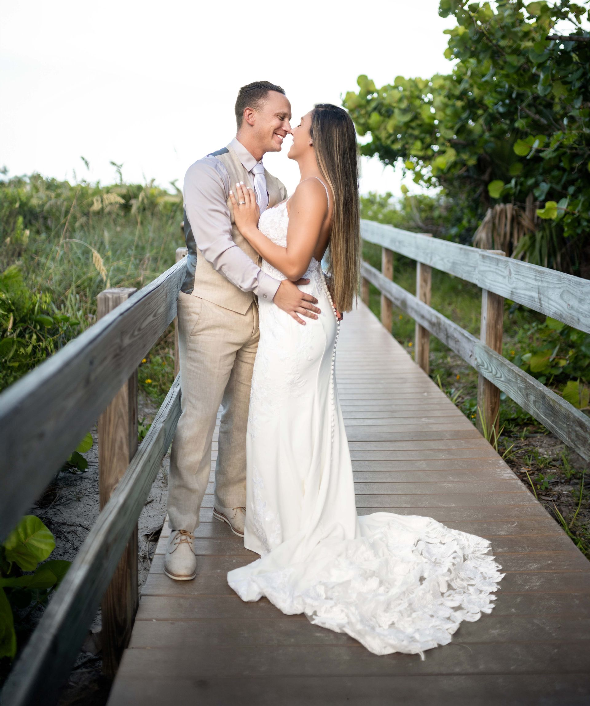 Bride and groom embrace on wooden bridge; she wears a white dress with a train, he wears a light suit.