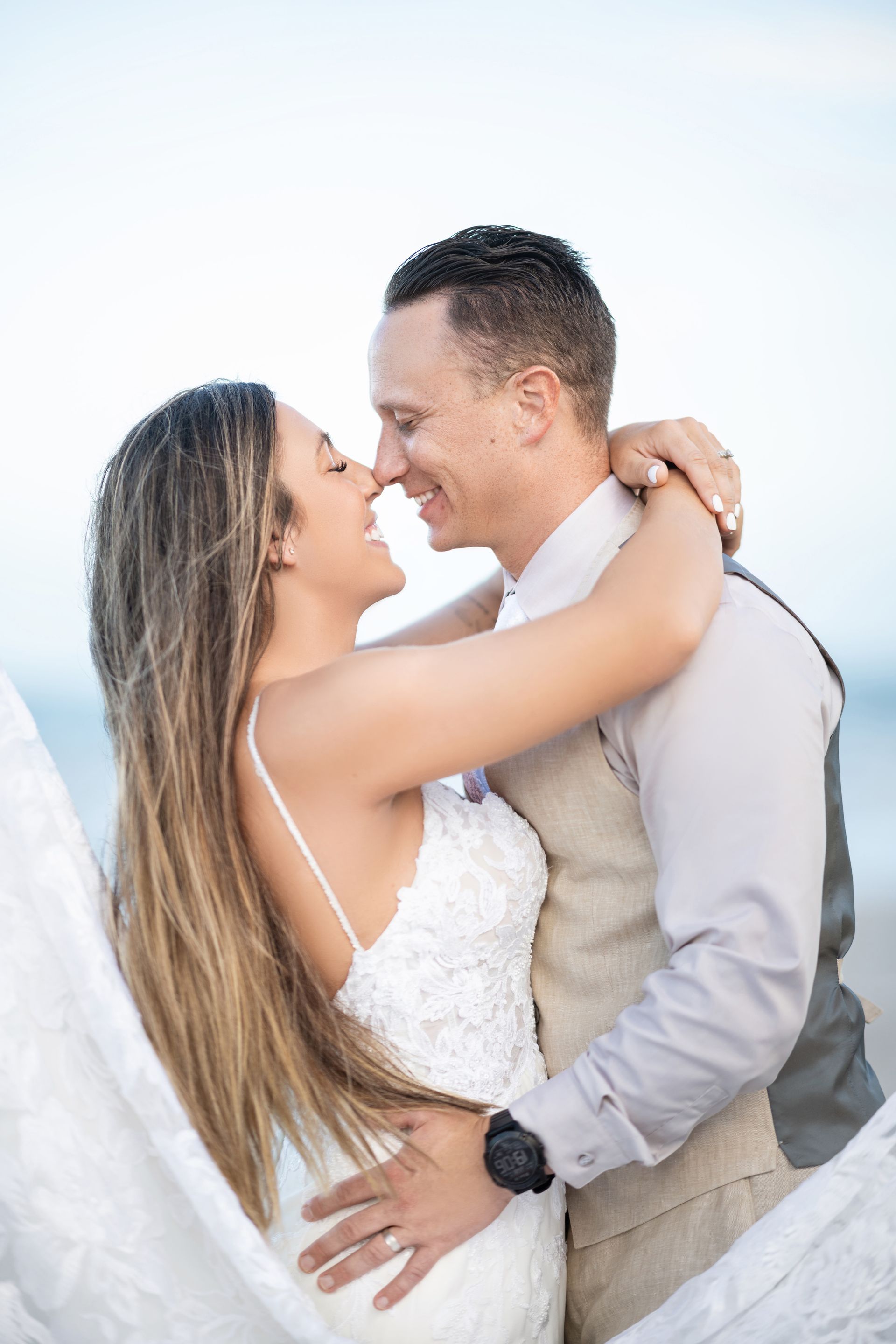 Bride and groom embrace on a beach, smiling and touching noses. Bride in white dress, groom in vest.