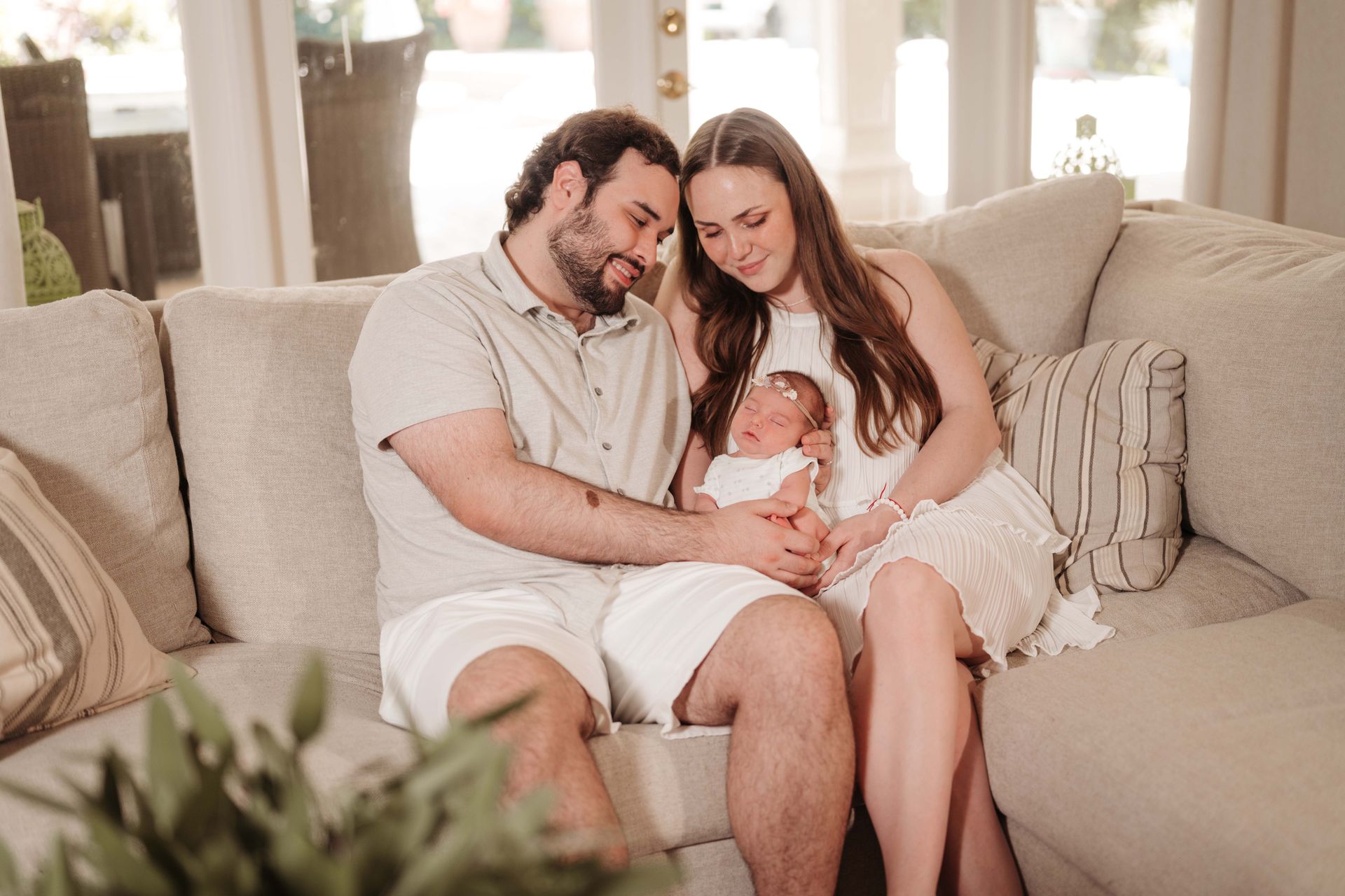 Parents on a couch hold their newborn baby; all are smiling. Sunny living room.