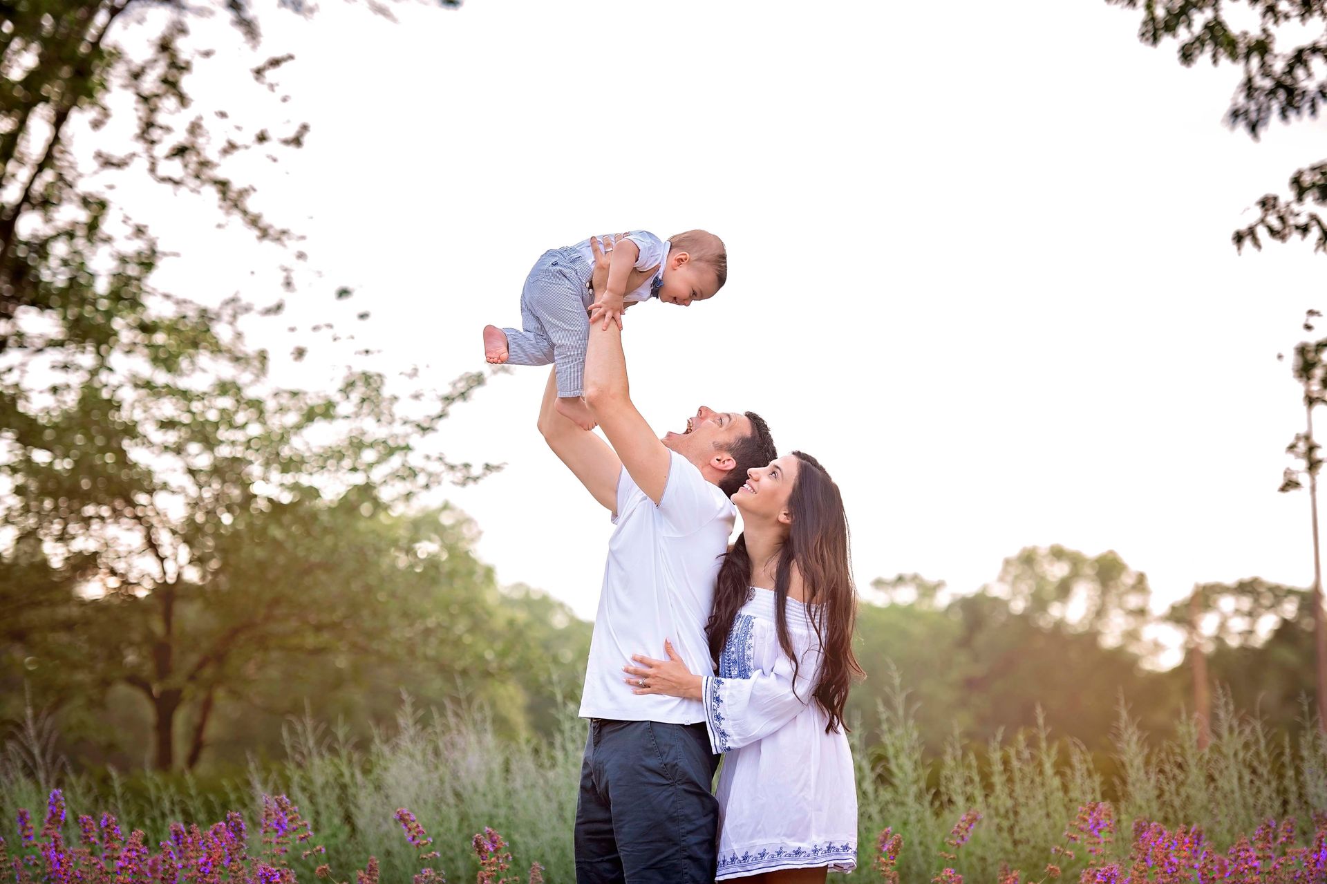 Family, dad holding up baby, mom watching. Purple flowers, green grass, and a bright sky.