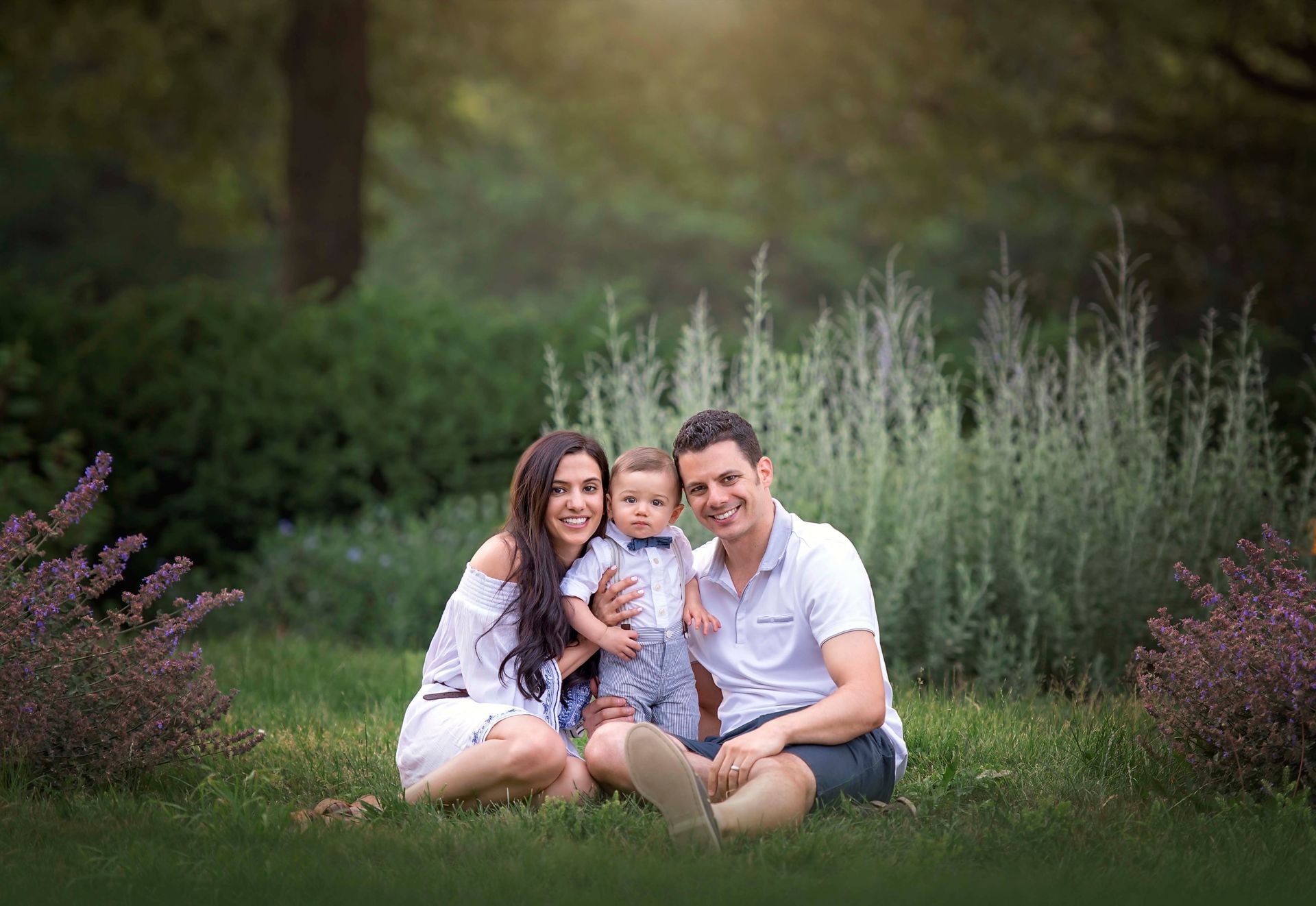 Family of three, sitting on grass. Smiling parents flank a small child in a field of green and purple, with a forest background.