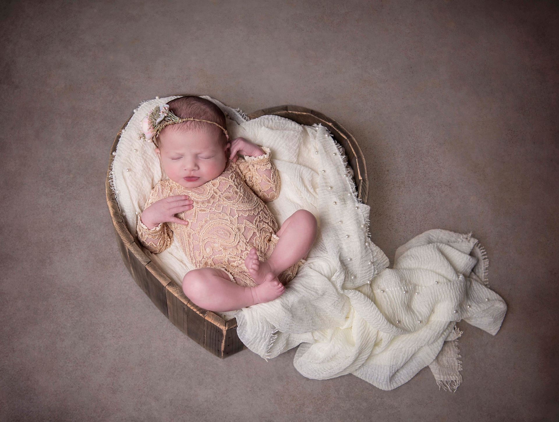 Newborn baby in a heart-shaped basket, wearing a tan outfit and headpiece, lying on a white blanket.