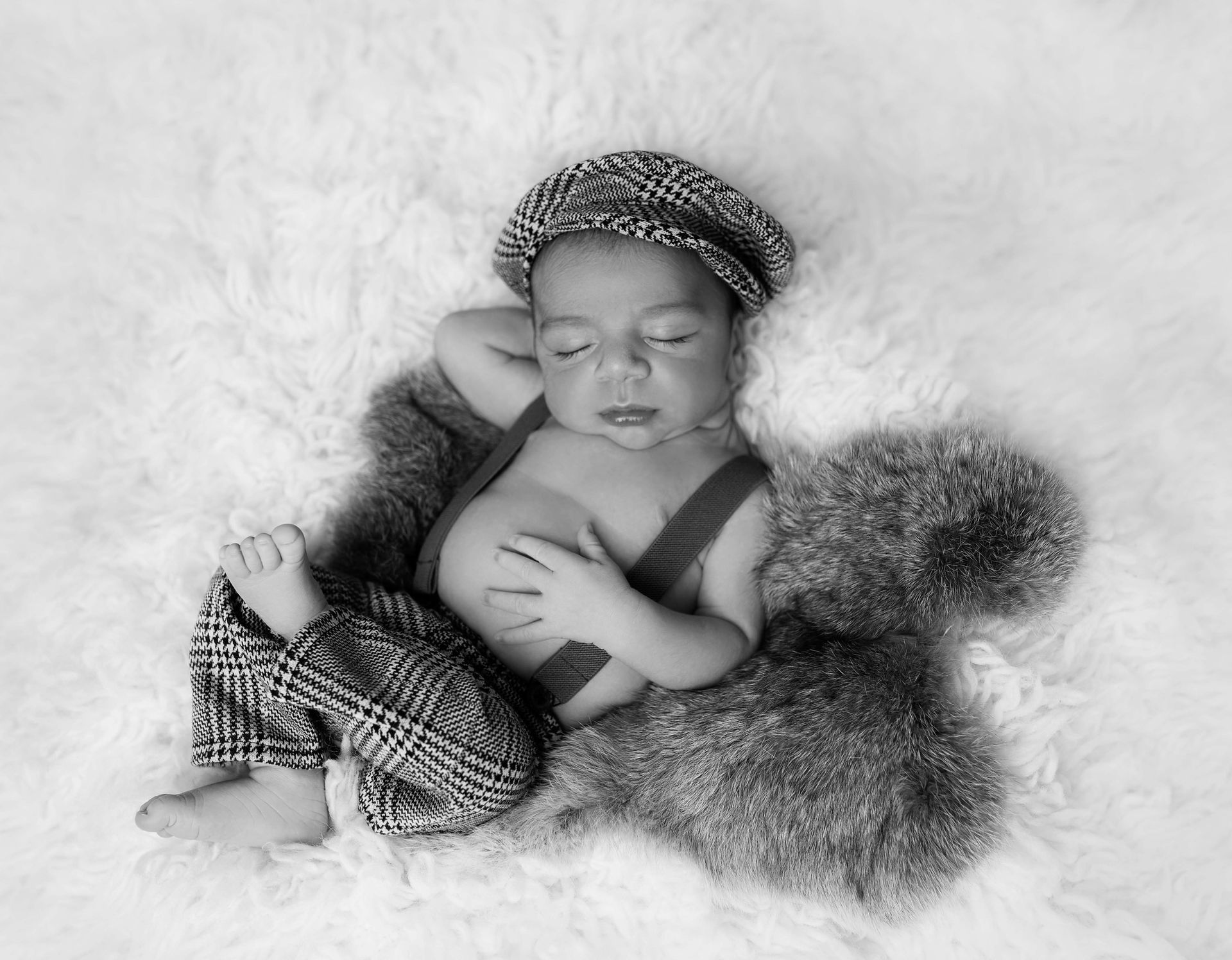 Newborn baby sleeping, wearing a plaid cap and pants with suspenders, on a furry rug.
