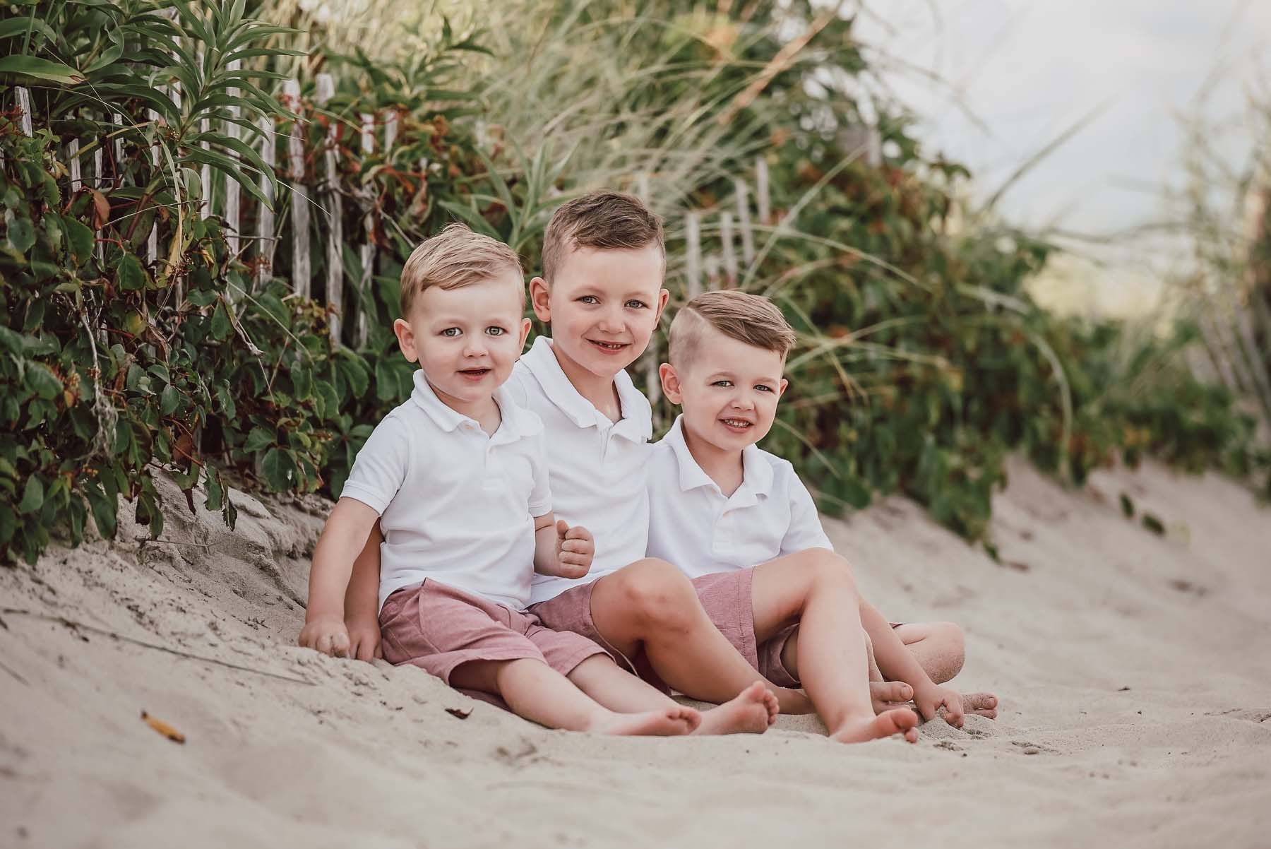Three young boys in white shirts and pink shorts sit on sand near a beach fence.