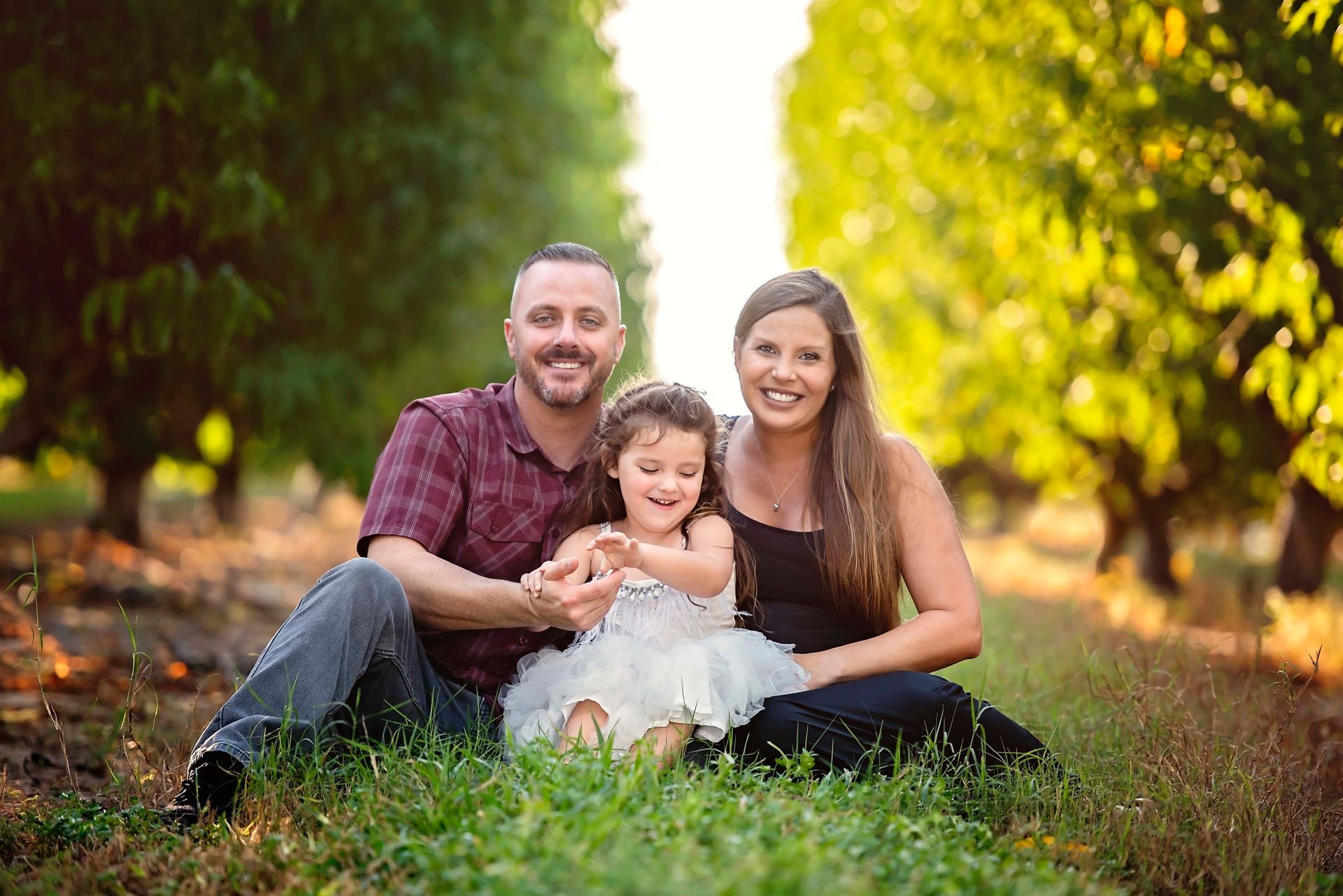 Family sitting in orchard: smiling parents with young daughter in white dress.