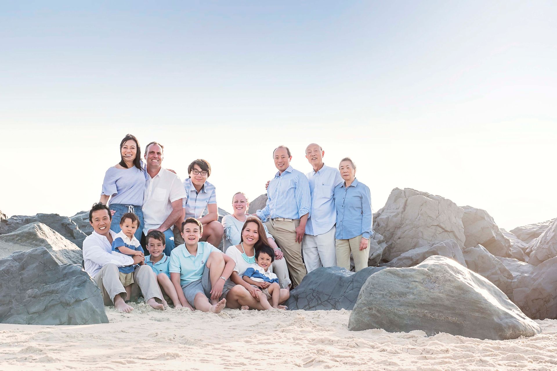 Large family posing on a beach, smiling. People of diverse ages and ethnicities wear light-colored clothing.