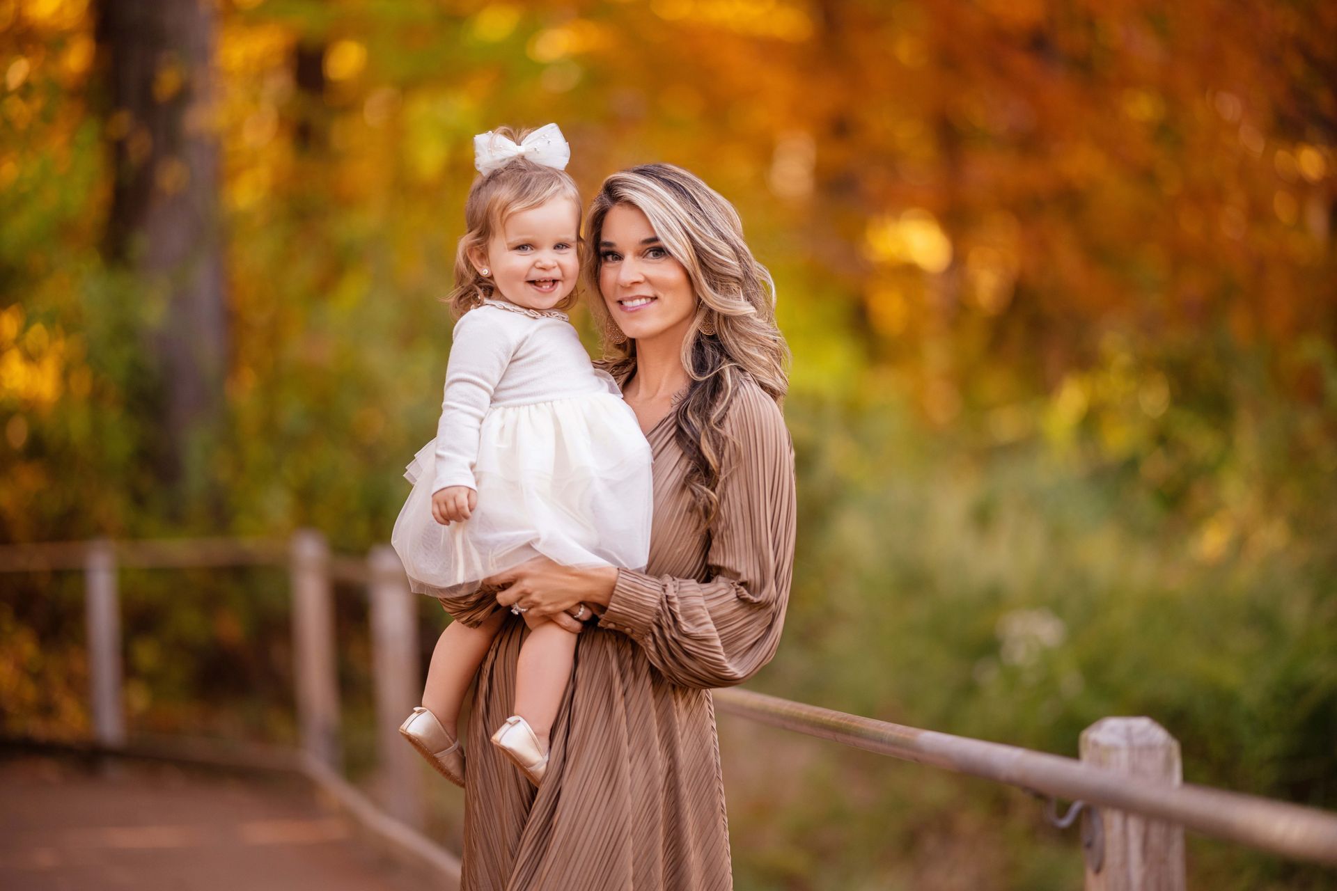 Woman holds a smiling toddler on a fall path. Both are smiling, surrounded by autumn colors.