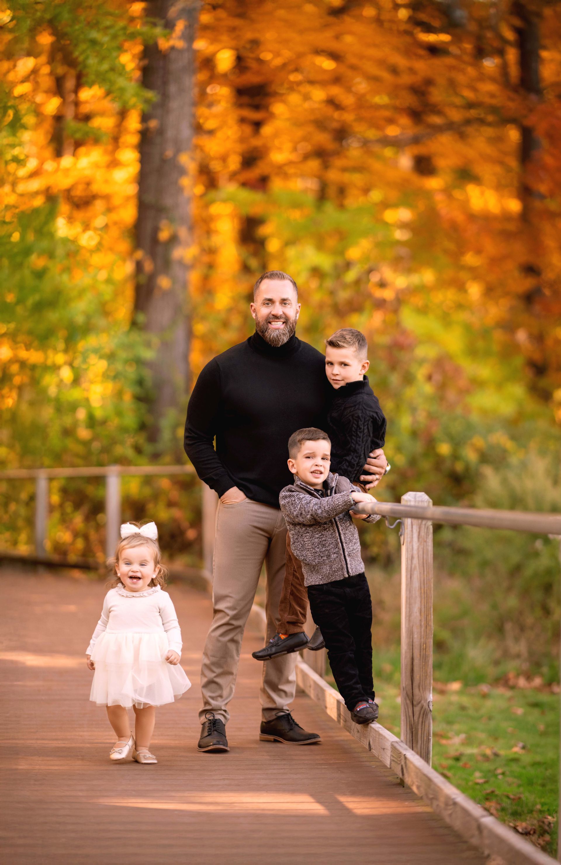 A father and three children pose on a wooden walkway in front of autumn foliage.