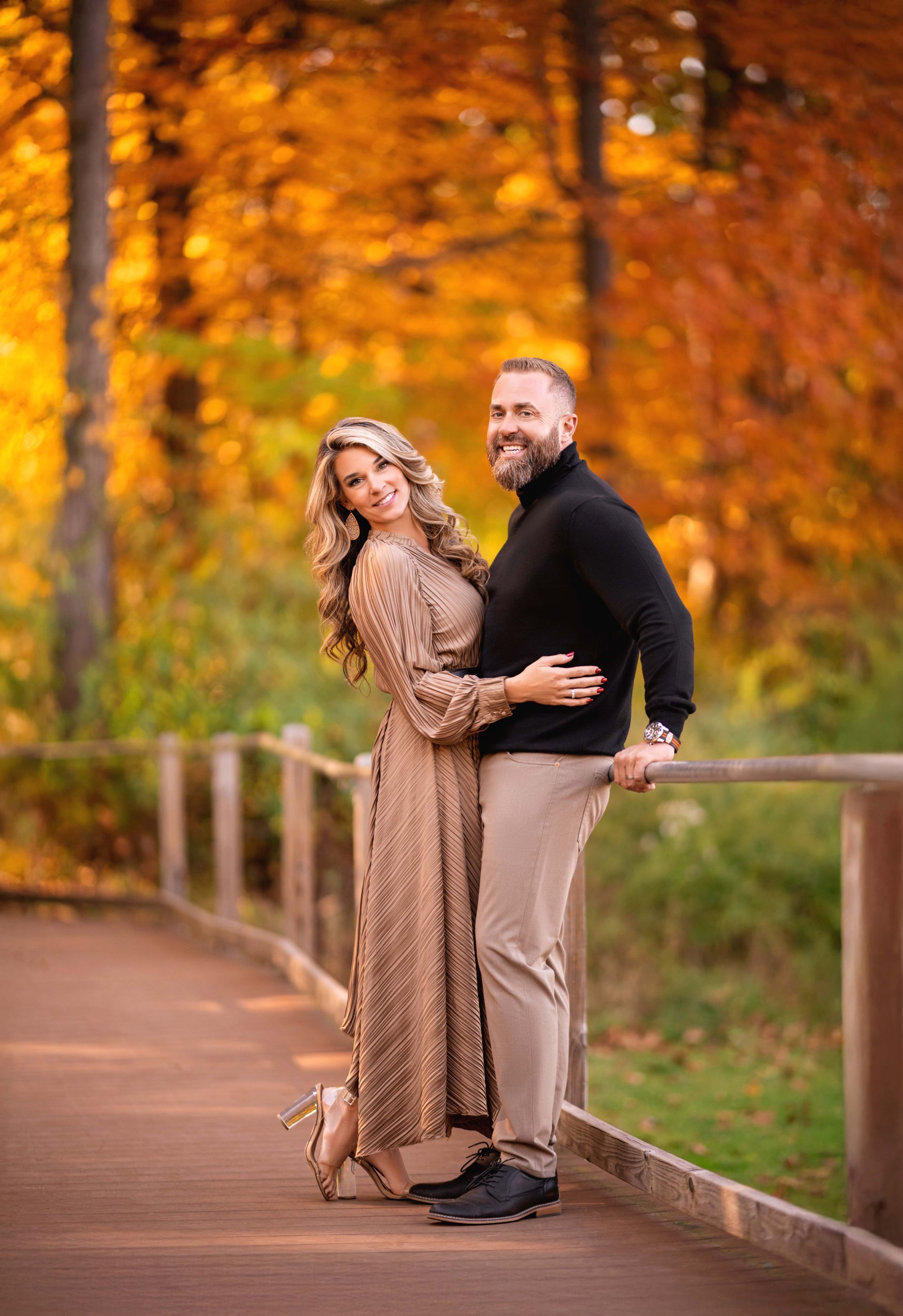 Couple in autumn scene, woman in patterned dress, man in black sweater, embracing on a wooden walkway with fall foliage.