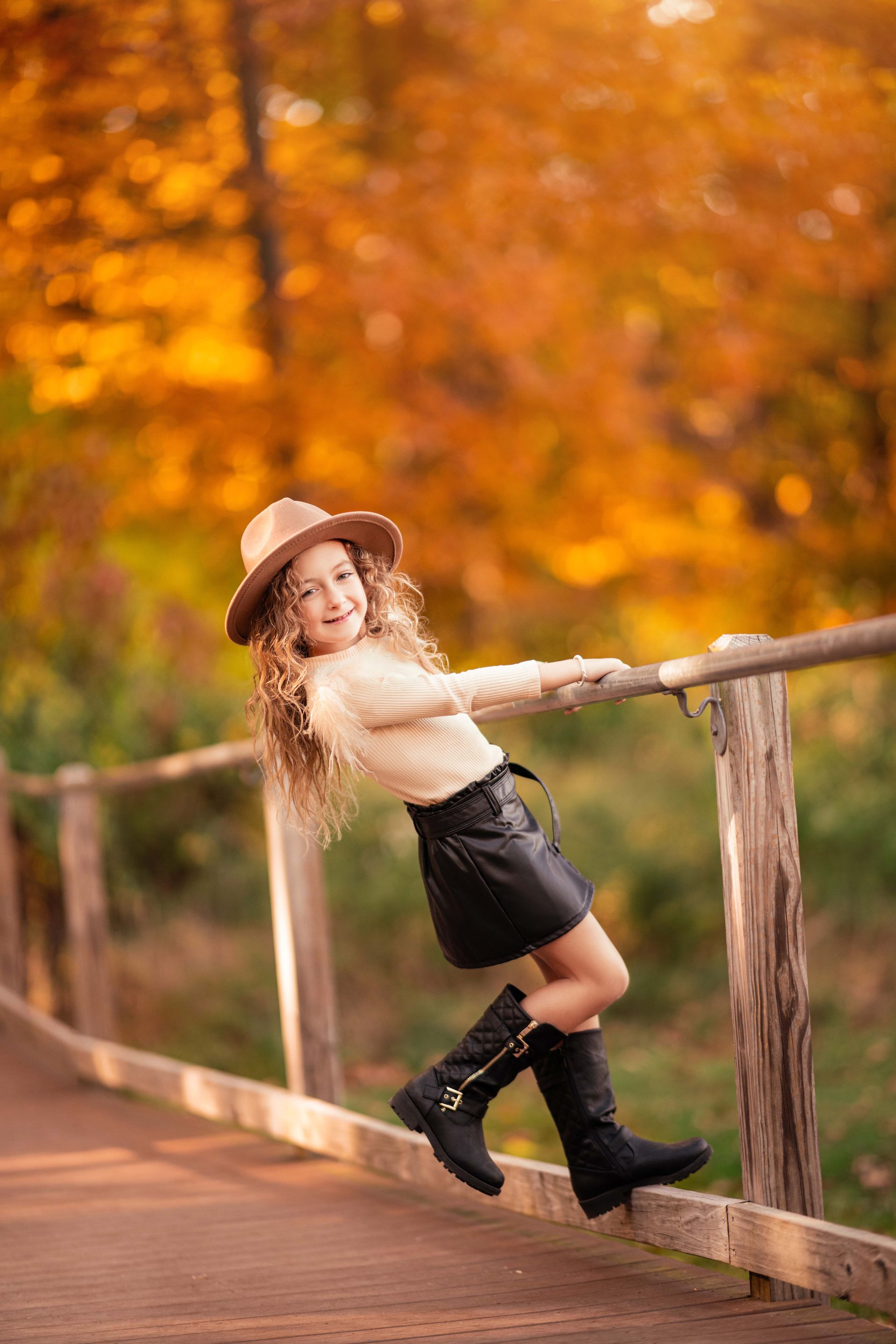 Girl in hat leans on wooden railing; fall foliage in background.