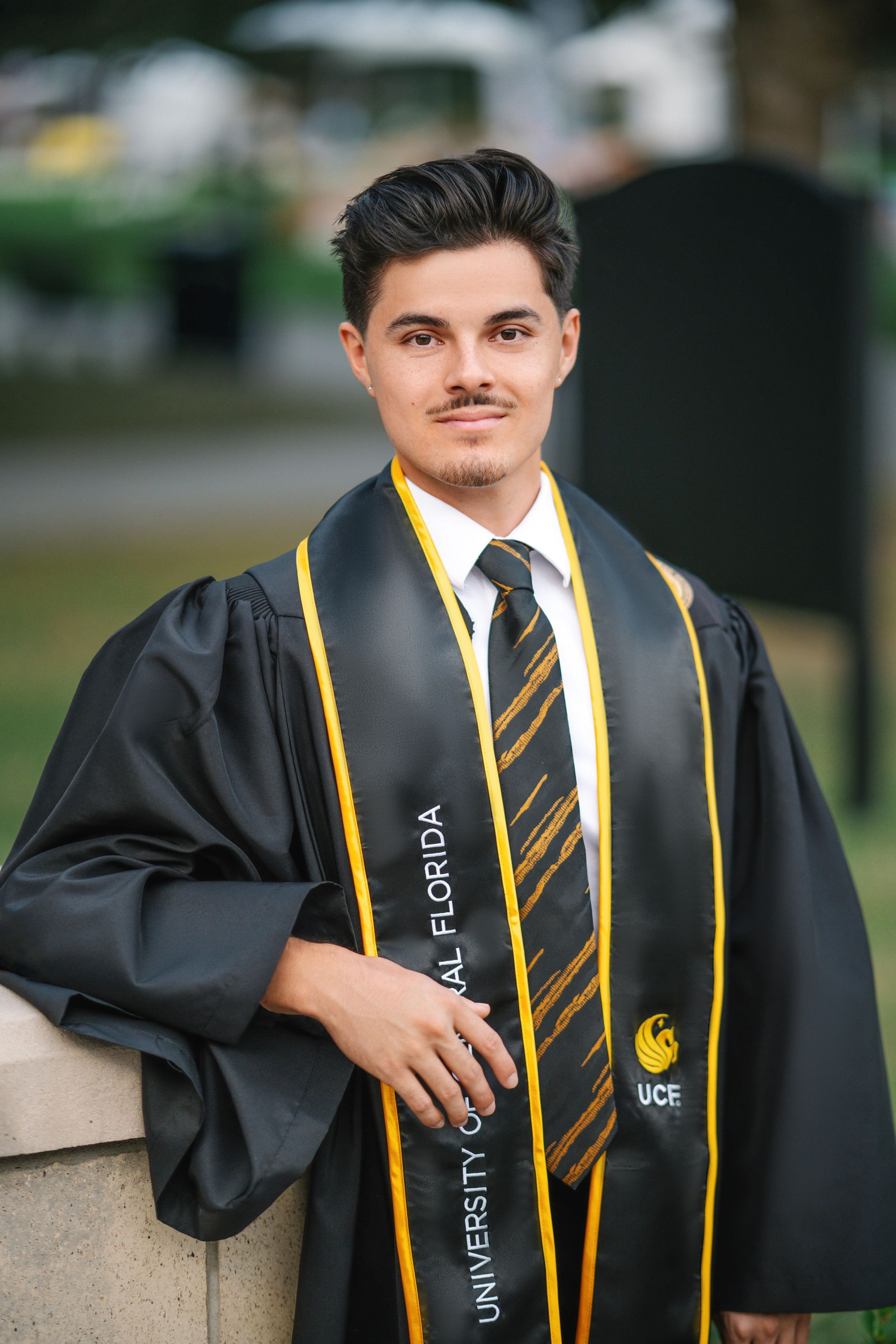 Woman in graduation cap and gown, smiling, outdoors on a wooden walkway.