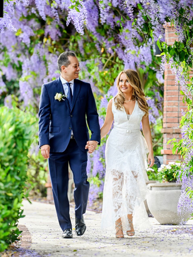 Bride and groom walking hand-in-hand under purple wisteria, brick wall on left, greenery on both sides.