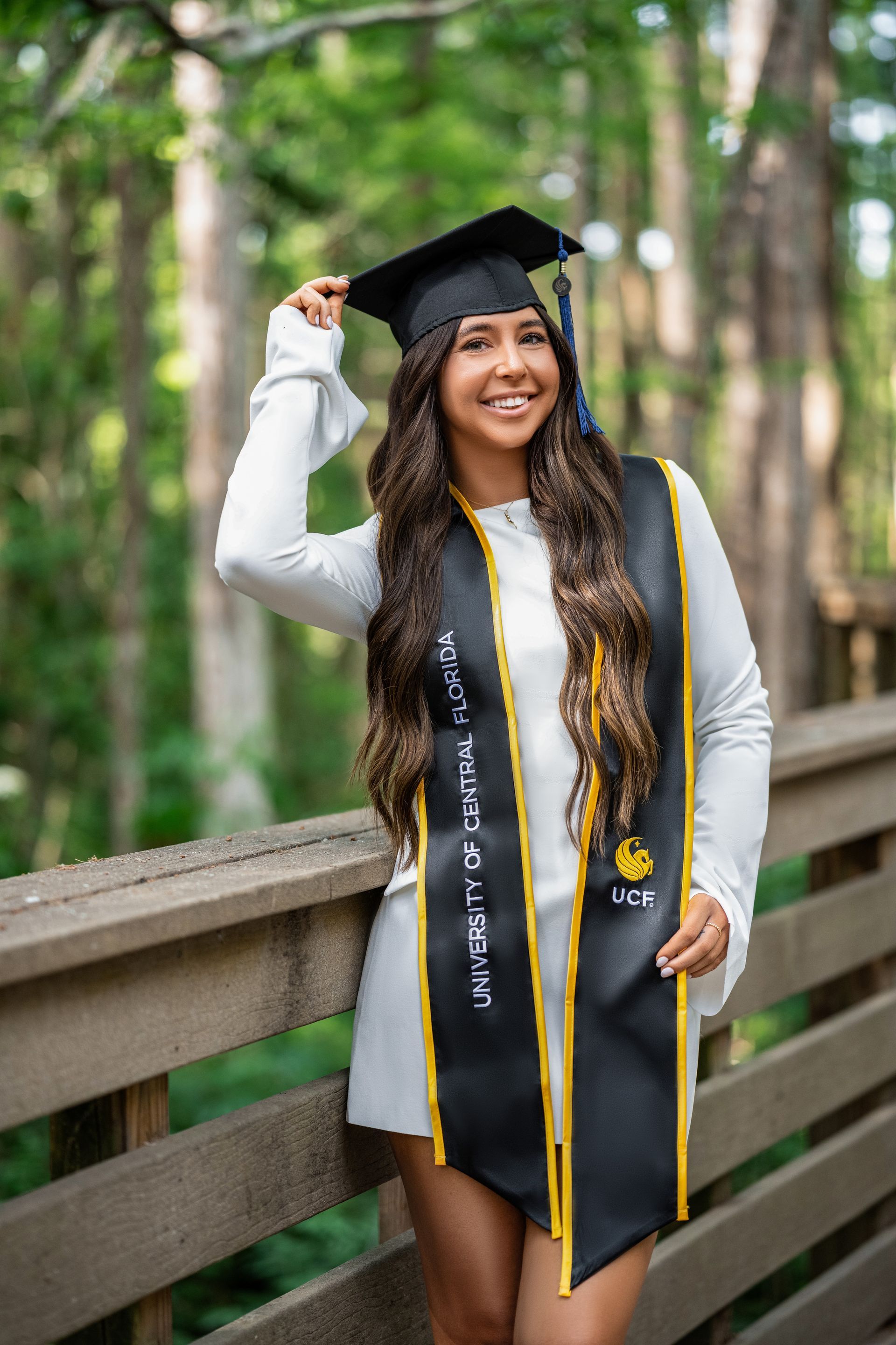 Woman in graduation cap and gown, smiling, outdoors on a wooden walkway.