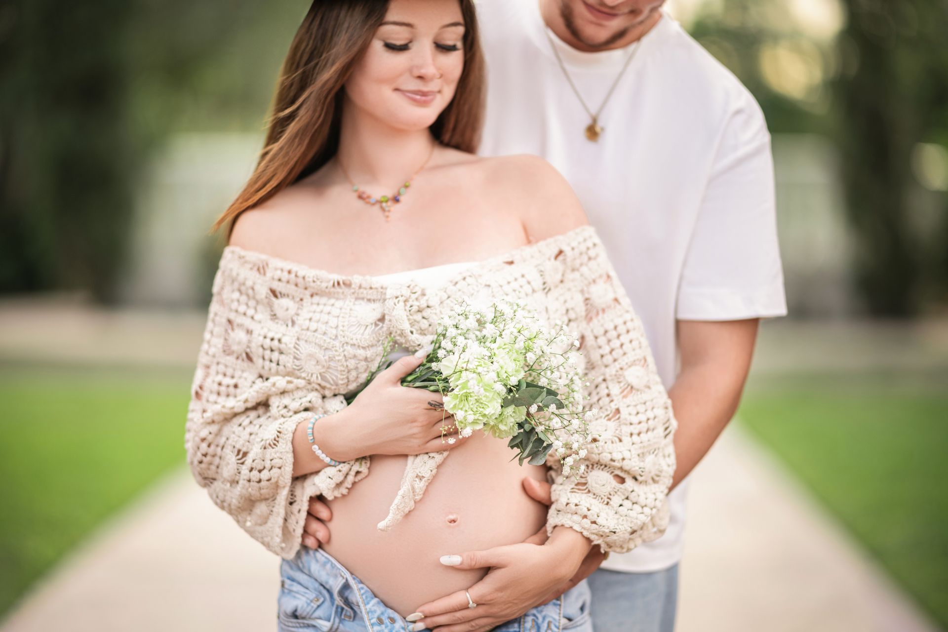 Pregnant woman and partner pose outdoors; she cradles her belly, holding flowers; he embraces her.