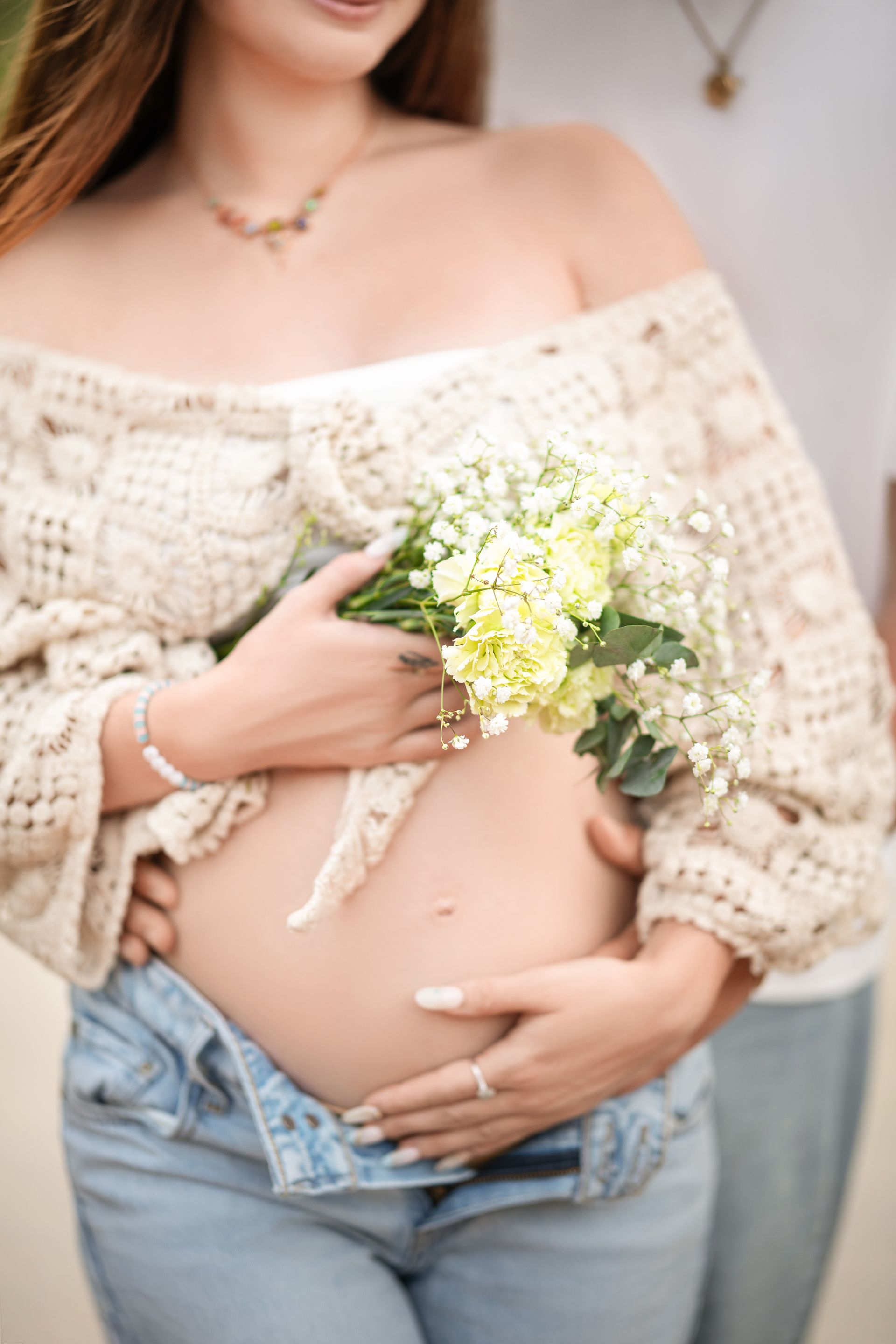 Pregnant woman in off-the-shoulder sweater, holding flowers, embracing partner; outdoors.