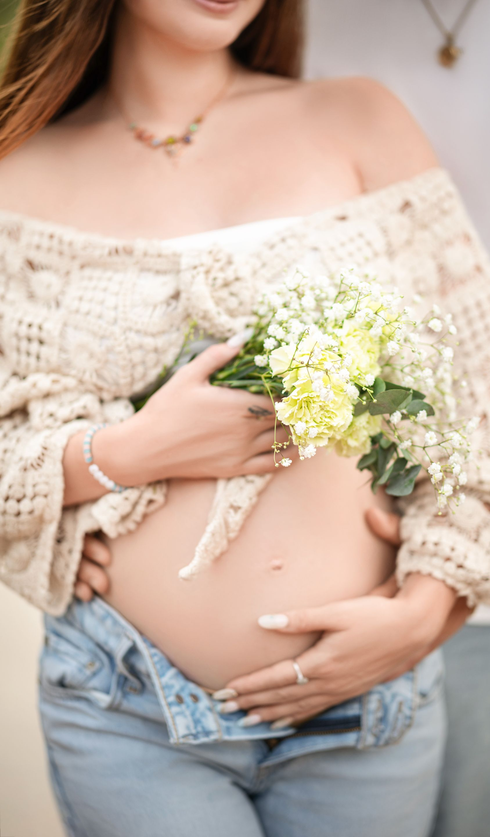 Pregnant woman holding flowers, belly exposed, wearing off-shoulder sweater and open jeans, embraced by someone.