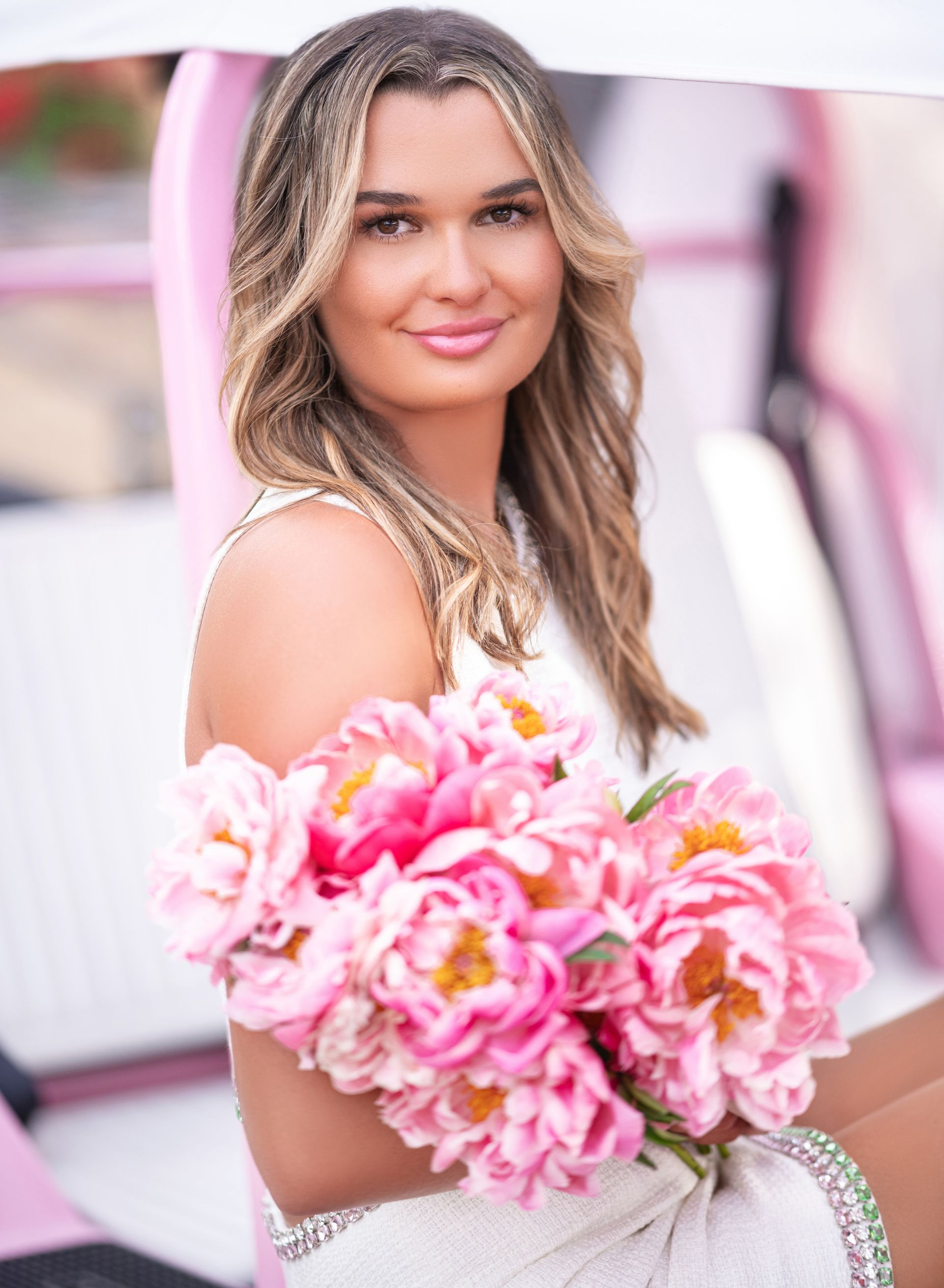 Woman with blonde hair holds pink peonies, sitting in a pink car.