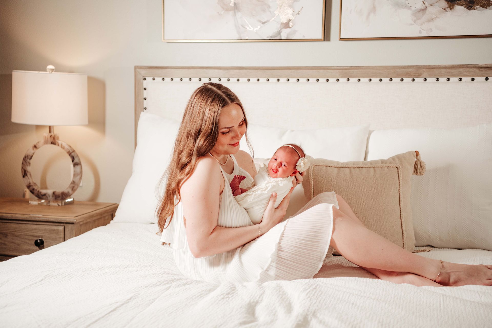 Mother in white dress, smiling, holding newborn wrapped in white, on a bed with neutral decor.