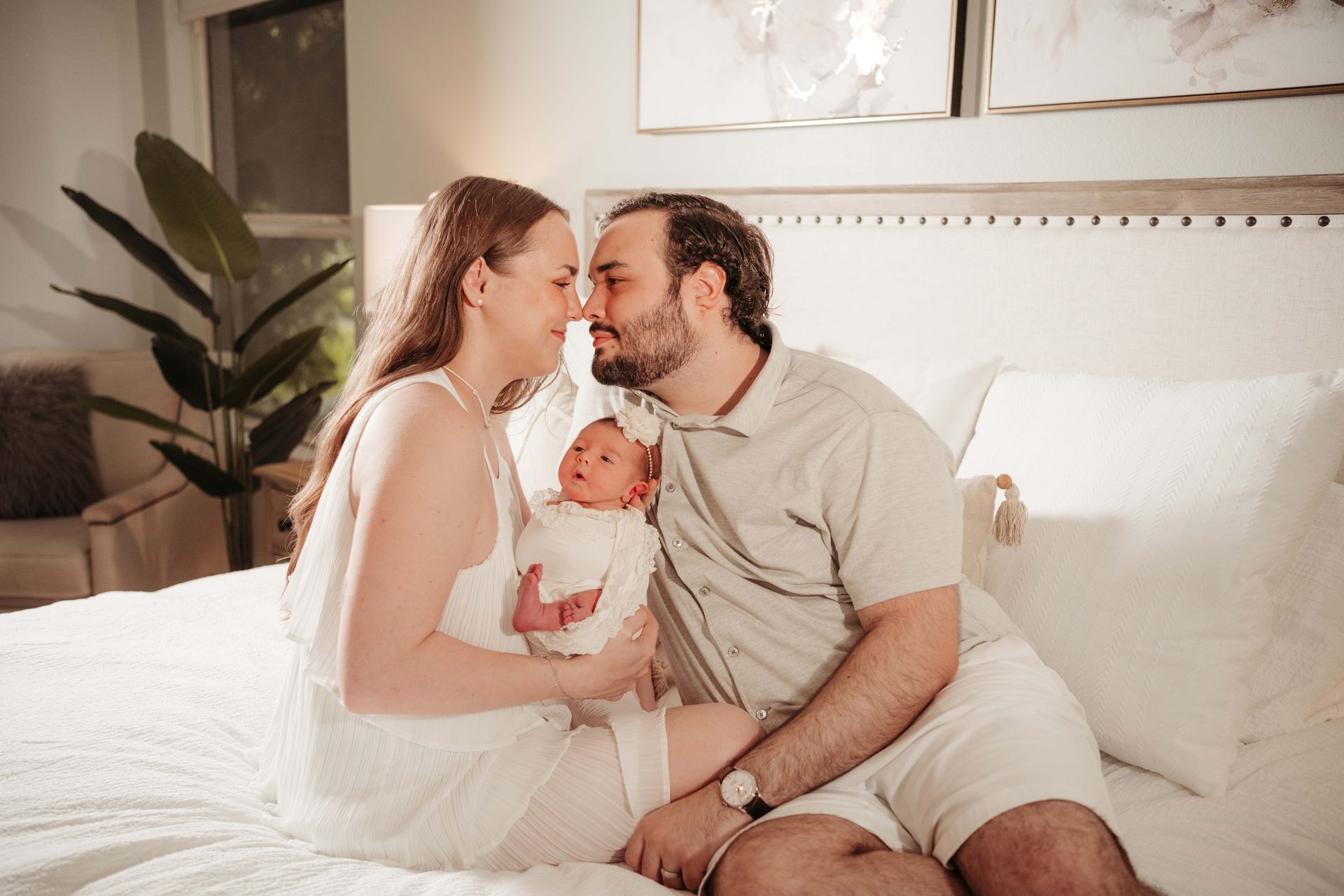 Parents in white clothing, touch foreheads with newborn baby on bed. Soft lighting, neutral tones.