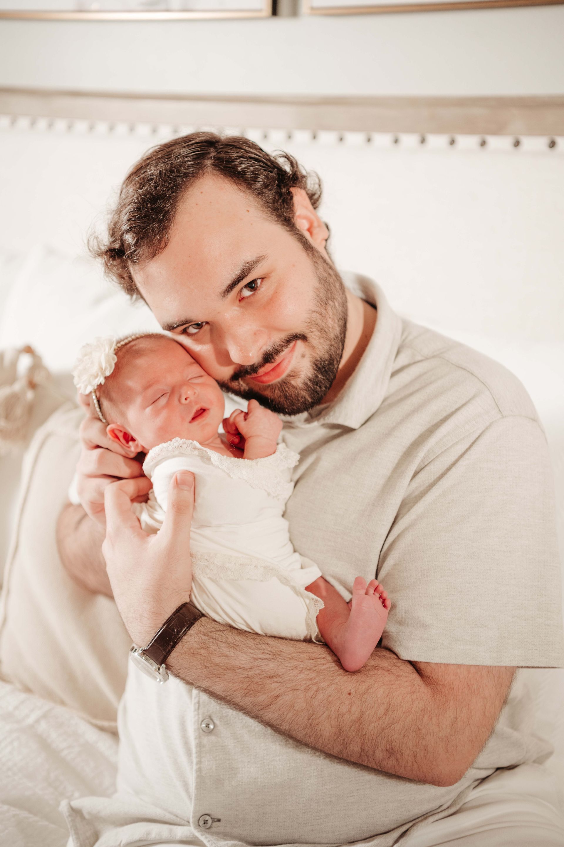 Man holding a newborn baby, both looking at the camera. Light skin, neutral tones, on a bed.