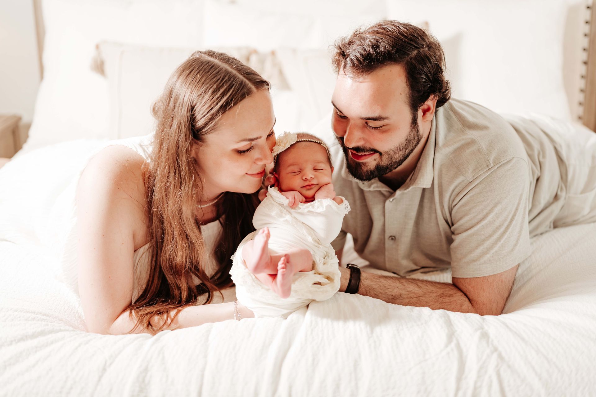 Parents gaze adoringly at newborn baby wrapped in white on a bed.
