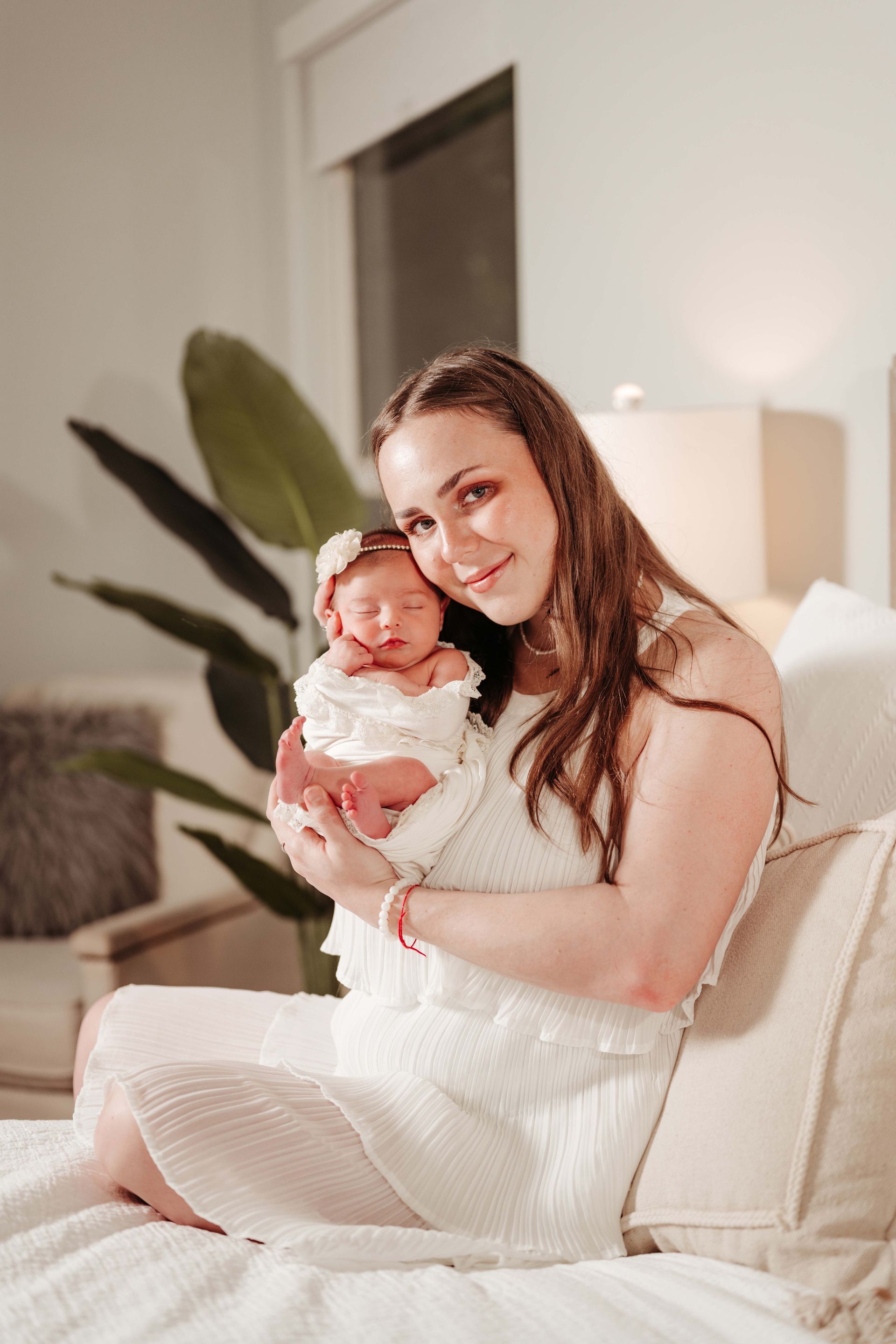 Woman in white dress holding newborn baby, both looking at camera, sitting on bed.
