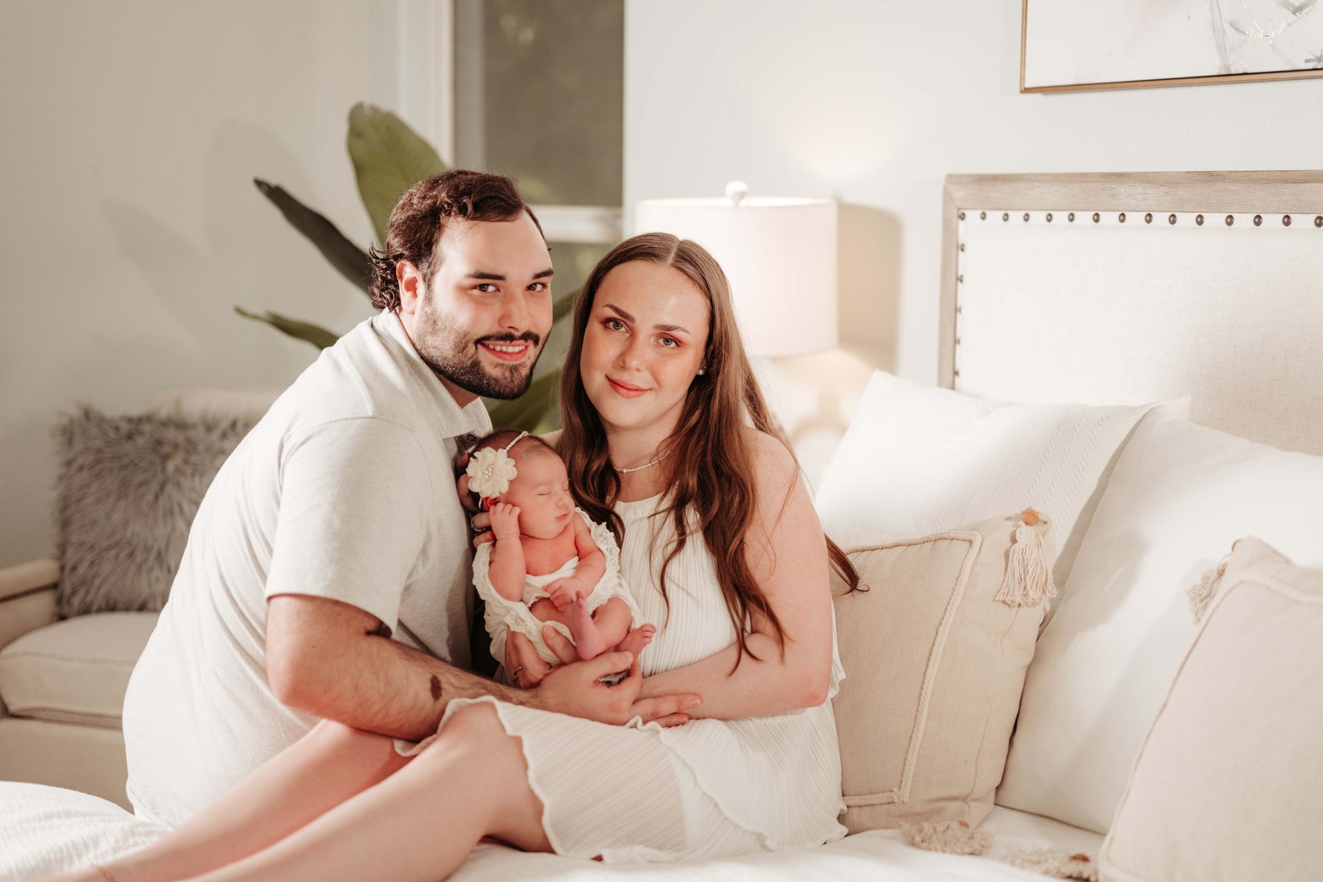 Parents holding newborn baby on bed. White outfits, neutral room, smiling.