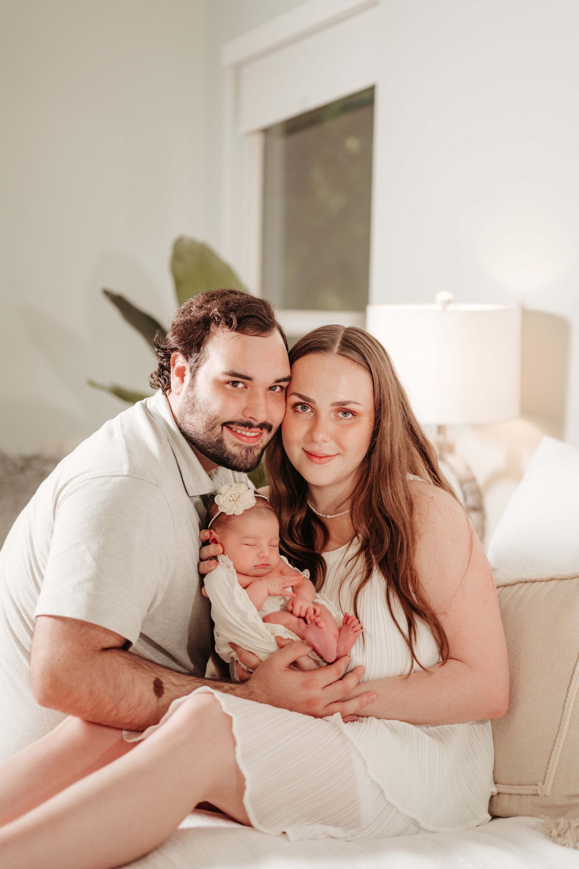 Family of three poses on a bed; parents hold newborn baby. Soft, natural light.