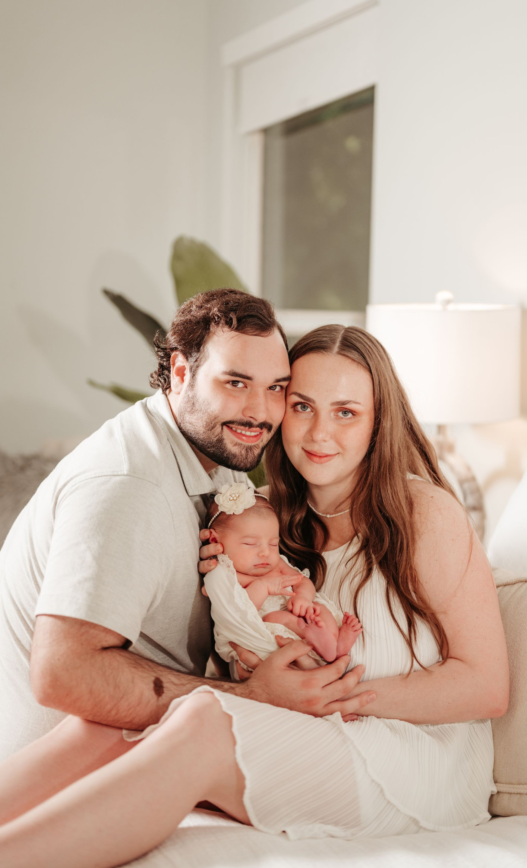 Parents smiling, holding newborn baby. Light-filled room. All wear white, family portrait.