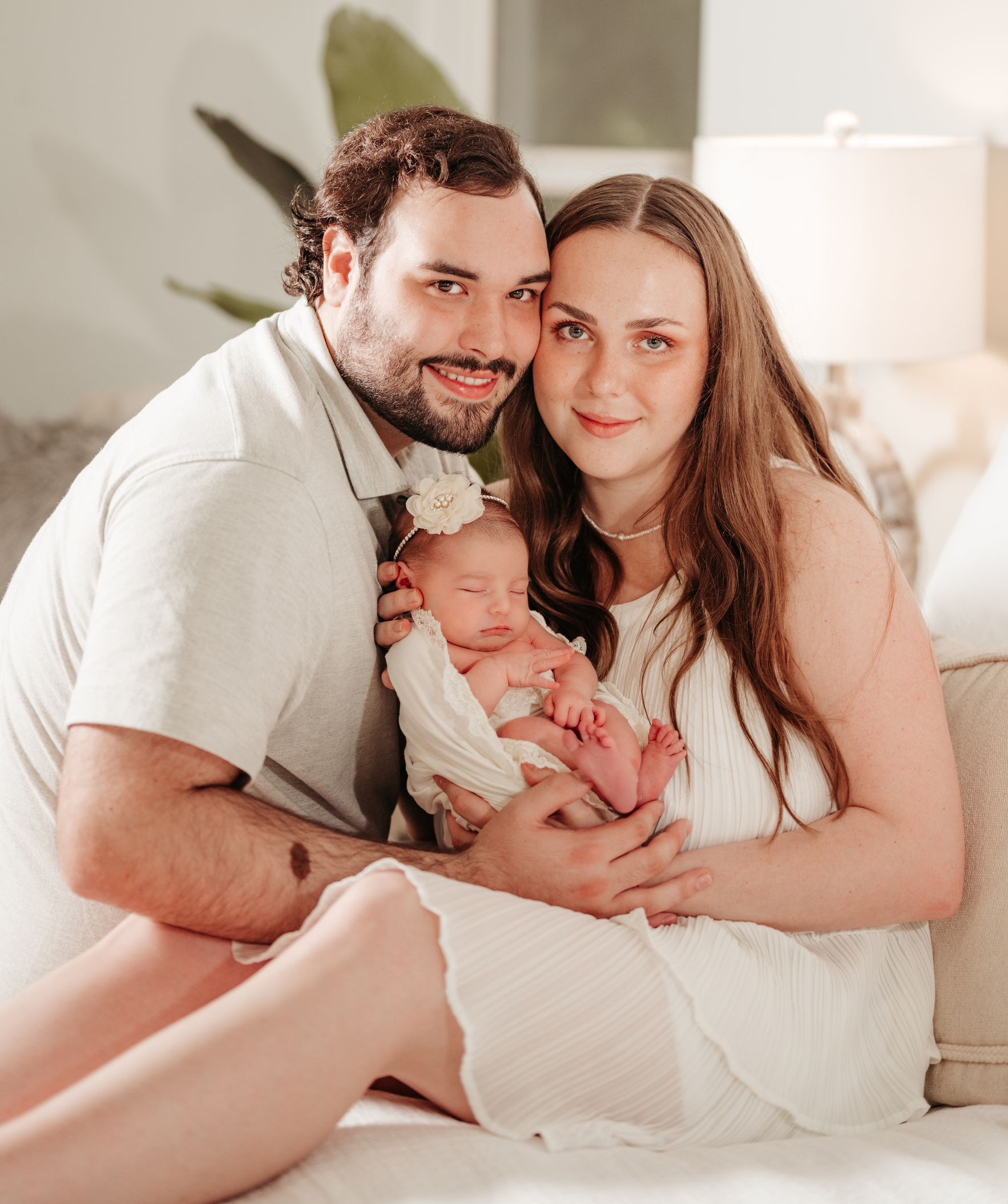 Parents cradle newborn, all in white, in a bright room.
