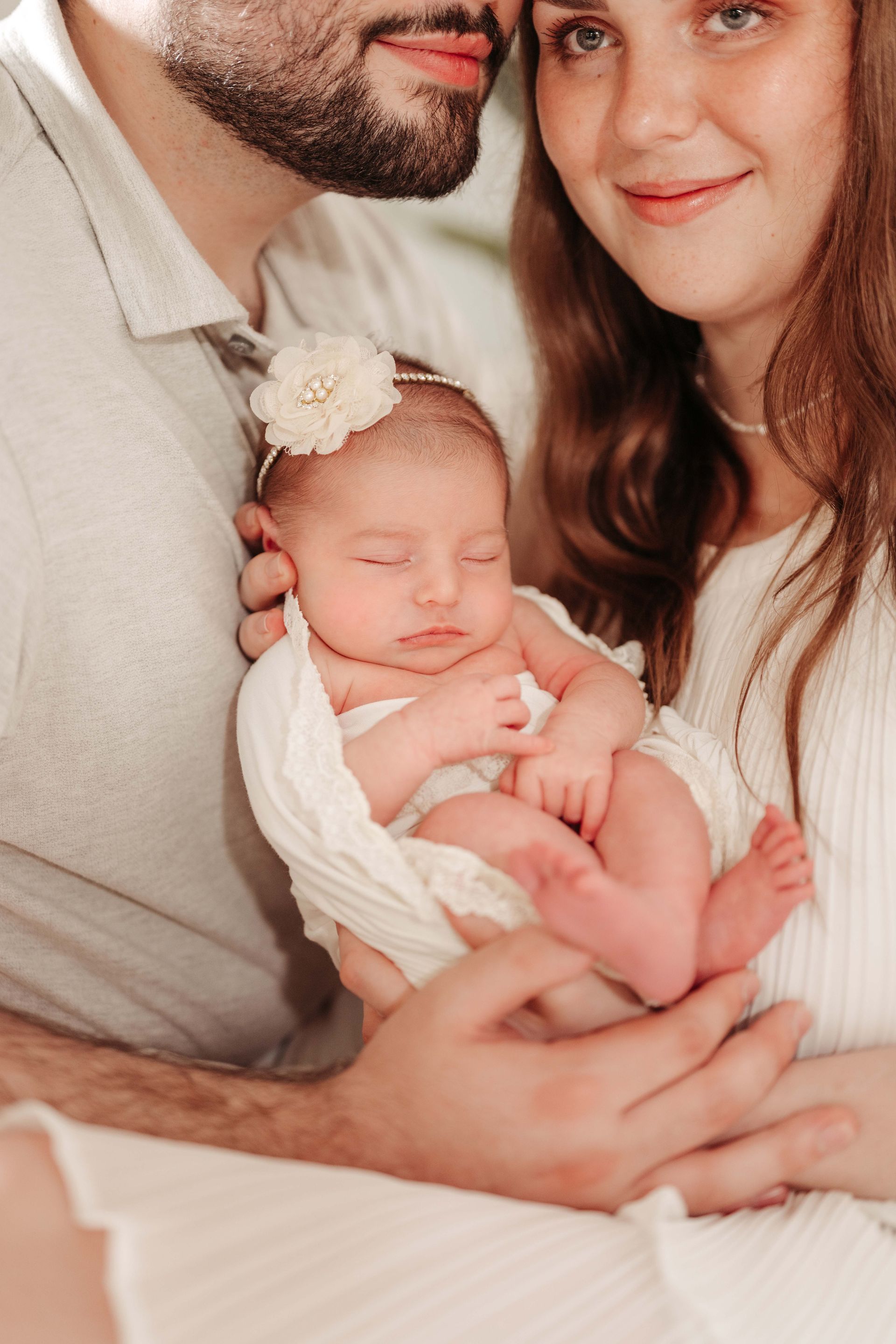 Parents holding a newborn baby, wrapped in white. The baby is sleeping, parents smiling.