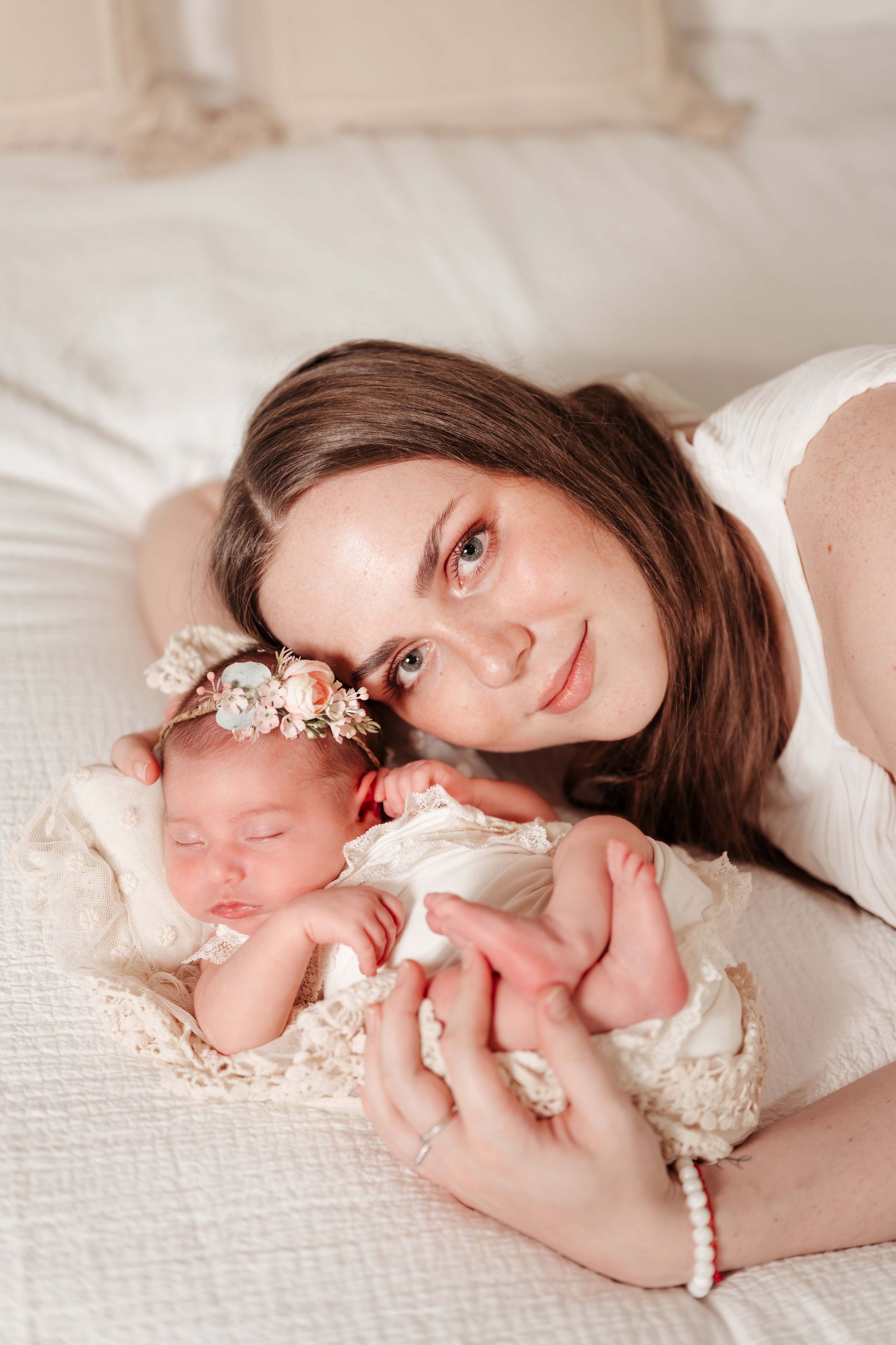 Mother smiles next to sleeping newborn in a lace basket on a white bed.