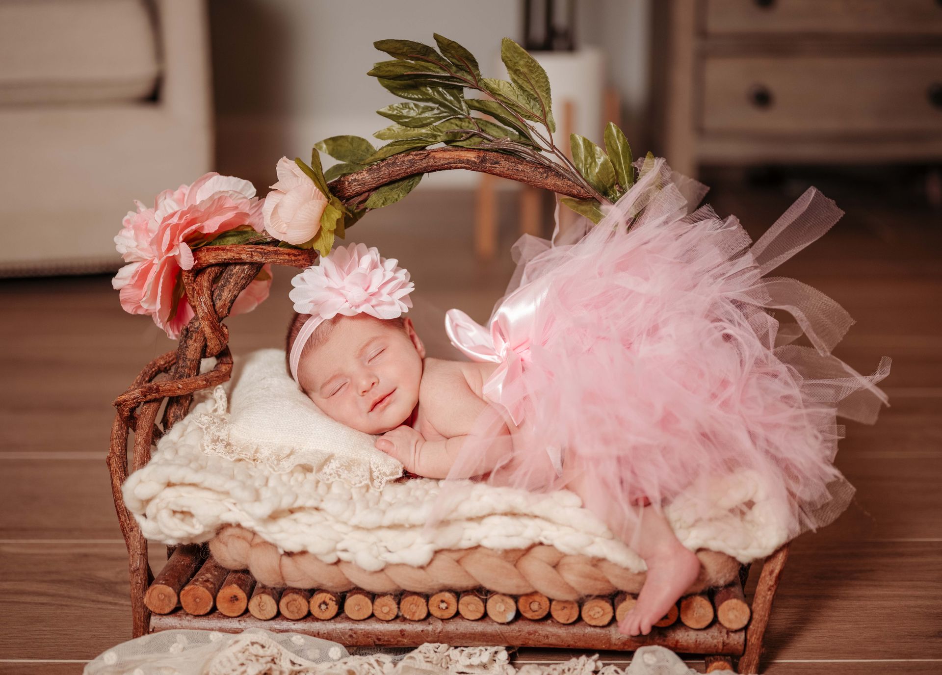 Newborn in a pink tutu and headband smiles while sleeping in a wooden bed decorated with flowers.