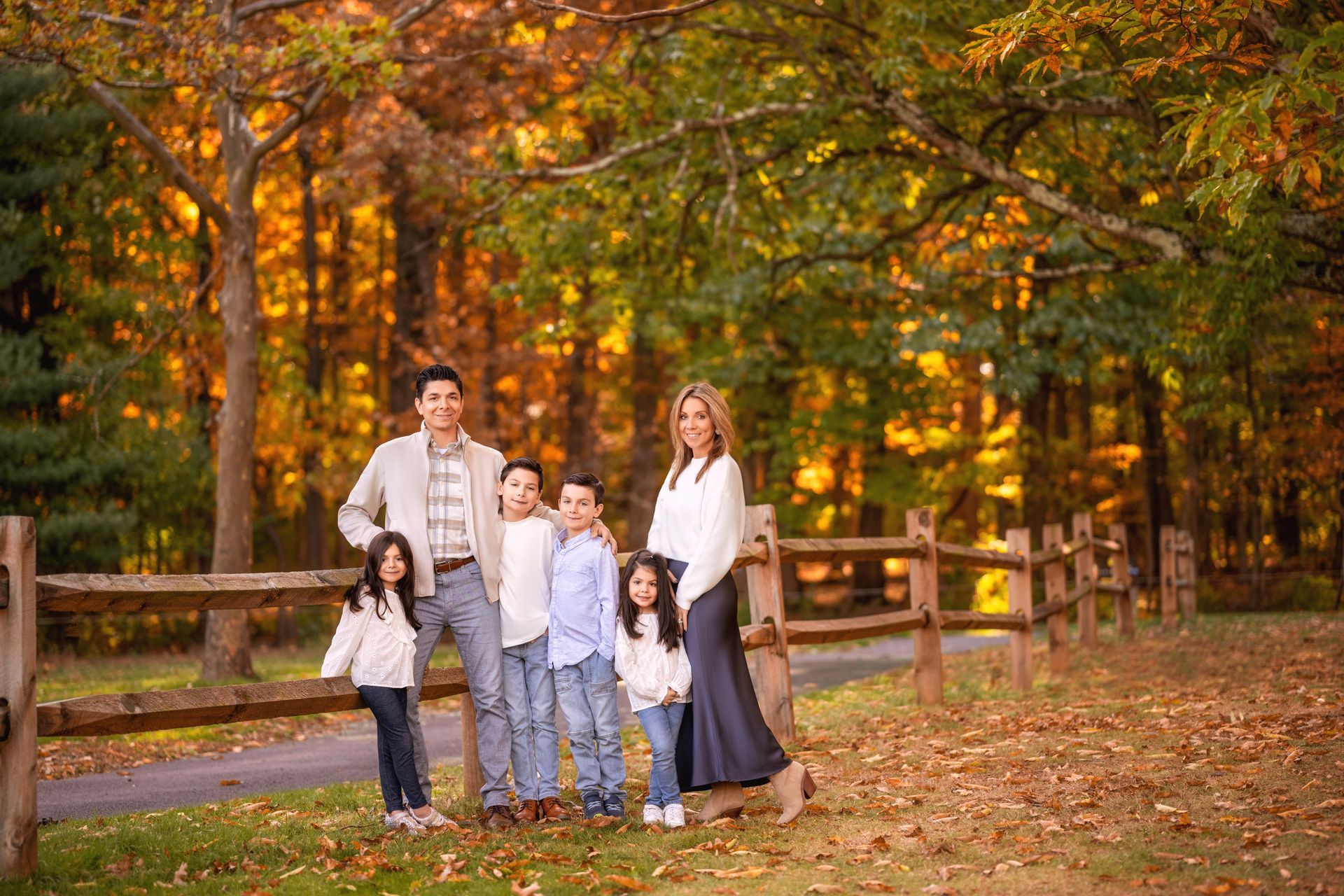 Family of six smiles in an autumn park, posed by a wooden fence.