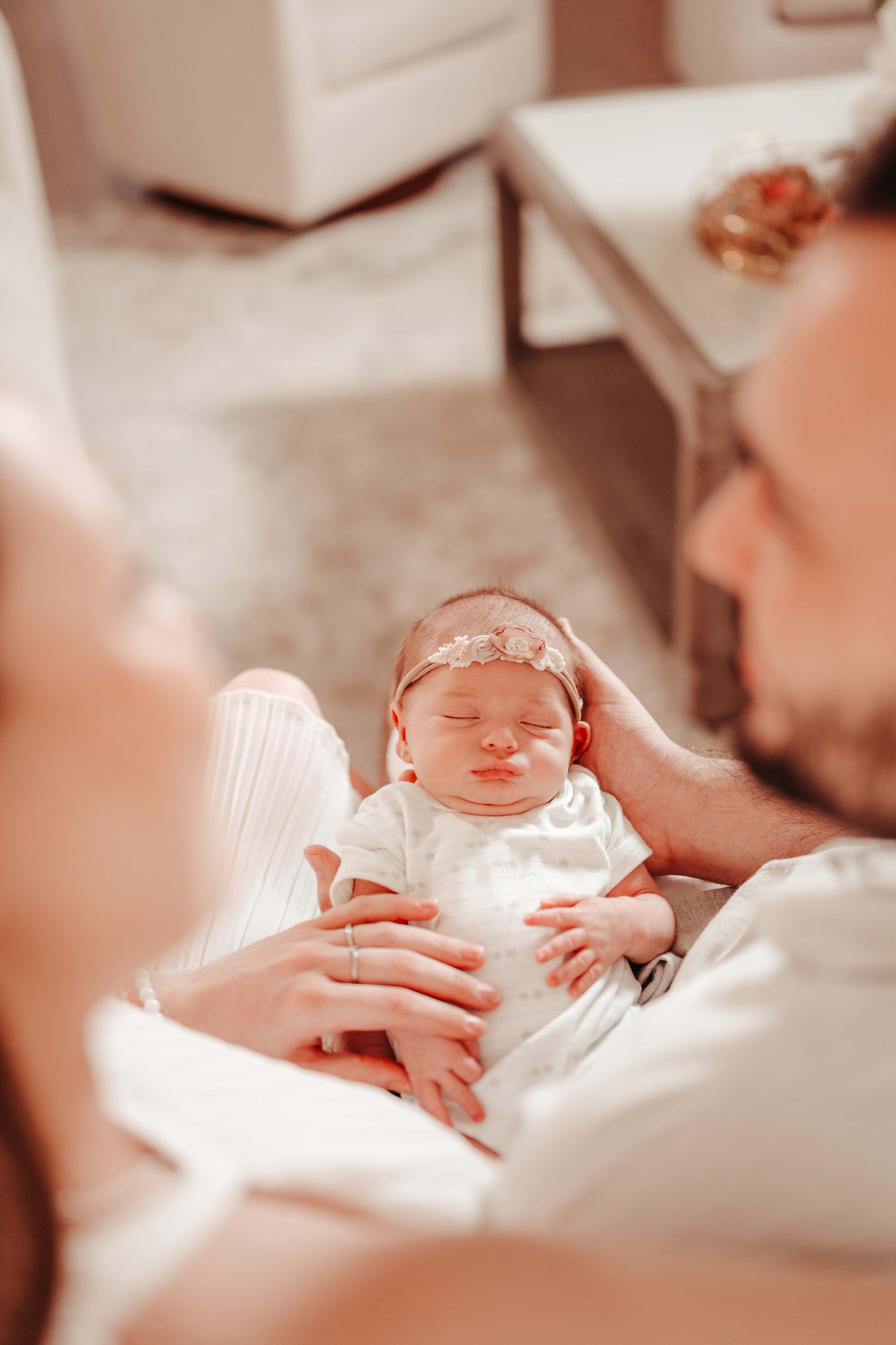 Parents holding a newborn baby wearing a floral headband and white onesie; soft lighting.