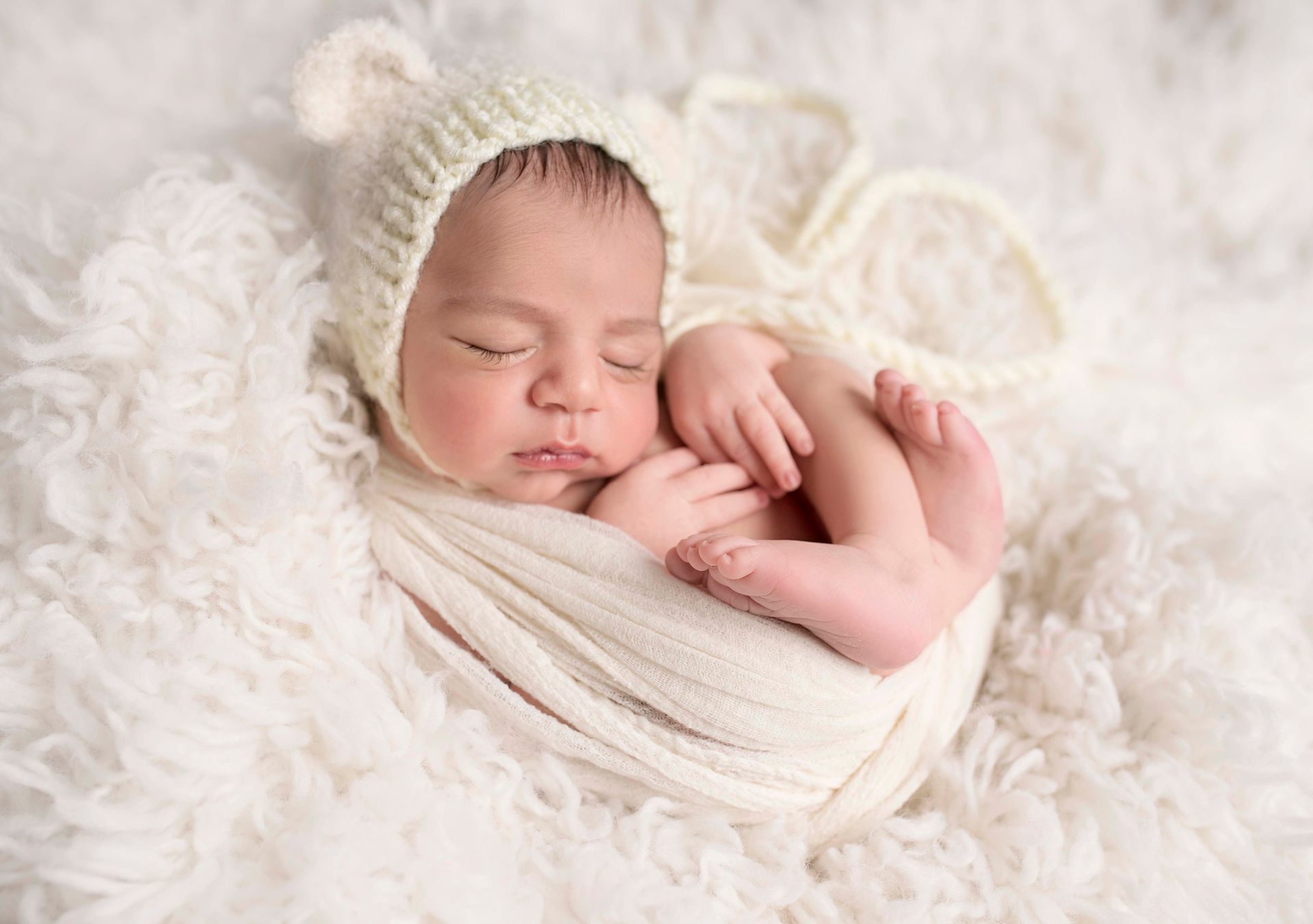 Newborn baby sleeping, wrapped in white blanket, wearing a cream bear hat on a fluffy white surface.