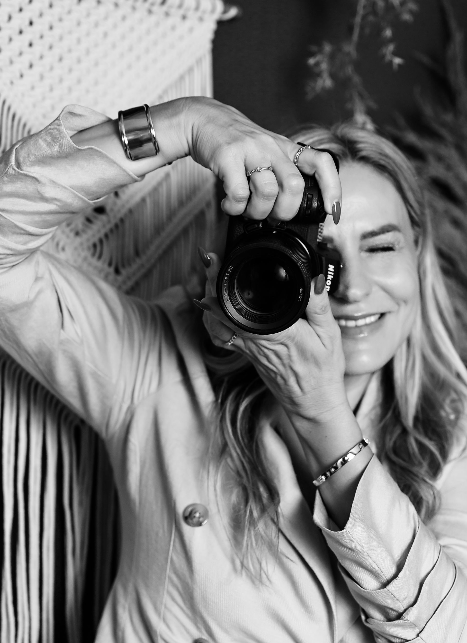 Blonde woman smiling while taking a photo with a camera, wearing jewelry. Black and white.