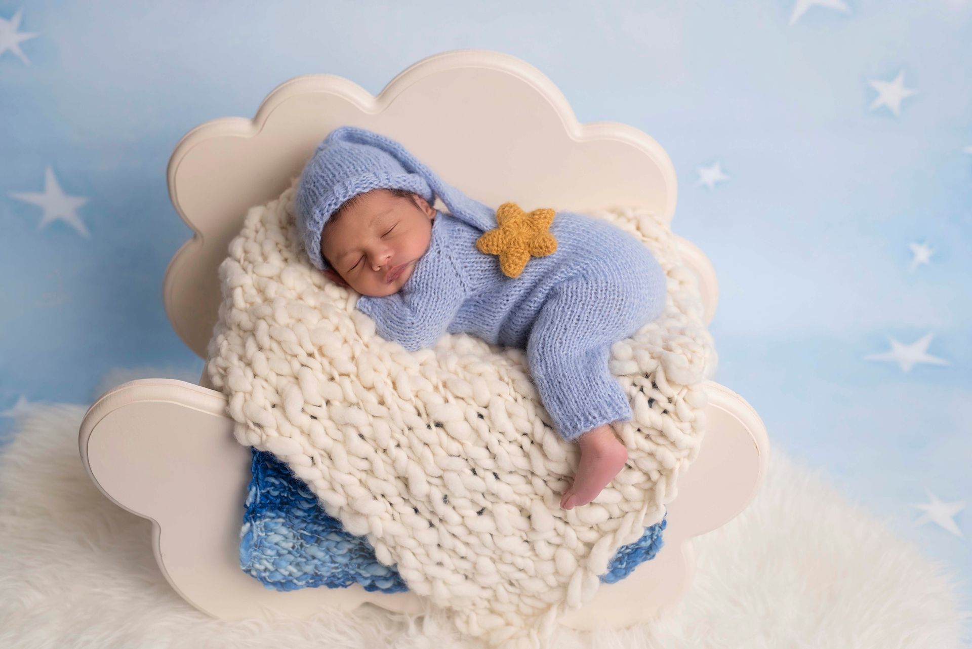 Newborn baby in blue knit outfit asleep on a cloud-shaped prop, with starry background.