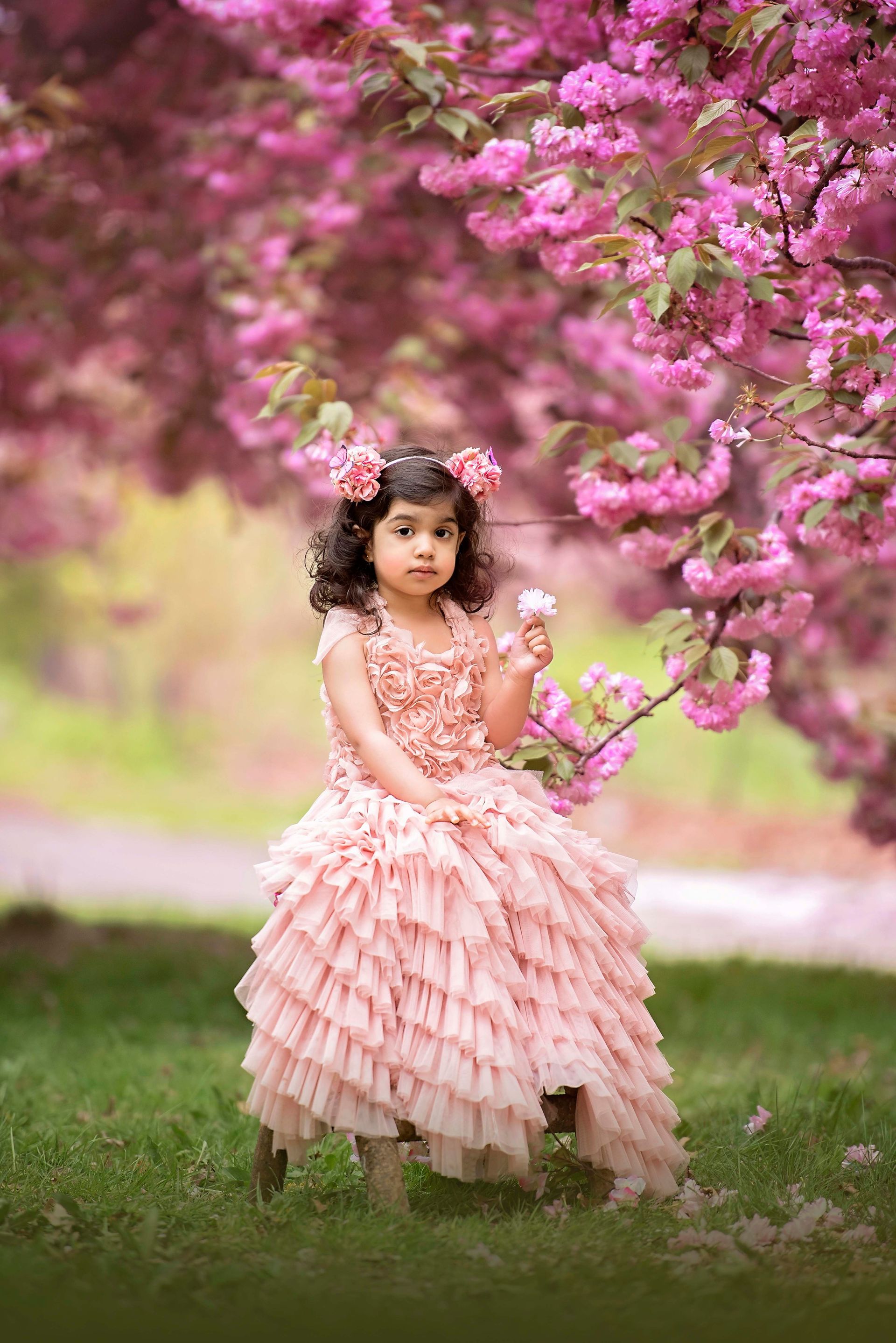 Girl in pink dress sits under flowering tree, holding a flower, looking at camera.