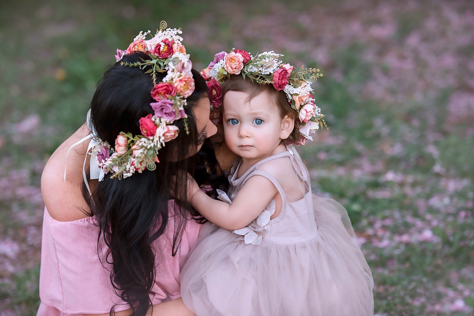 Mother and child wearing flower crowns, embracing outdoors, looking at the camera.