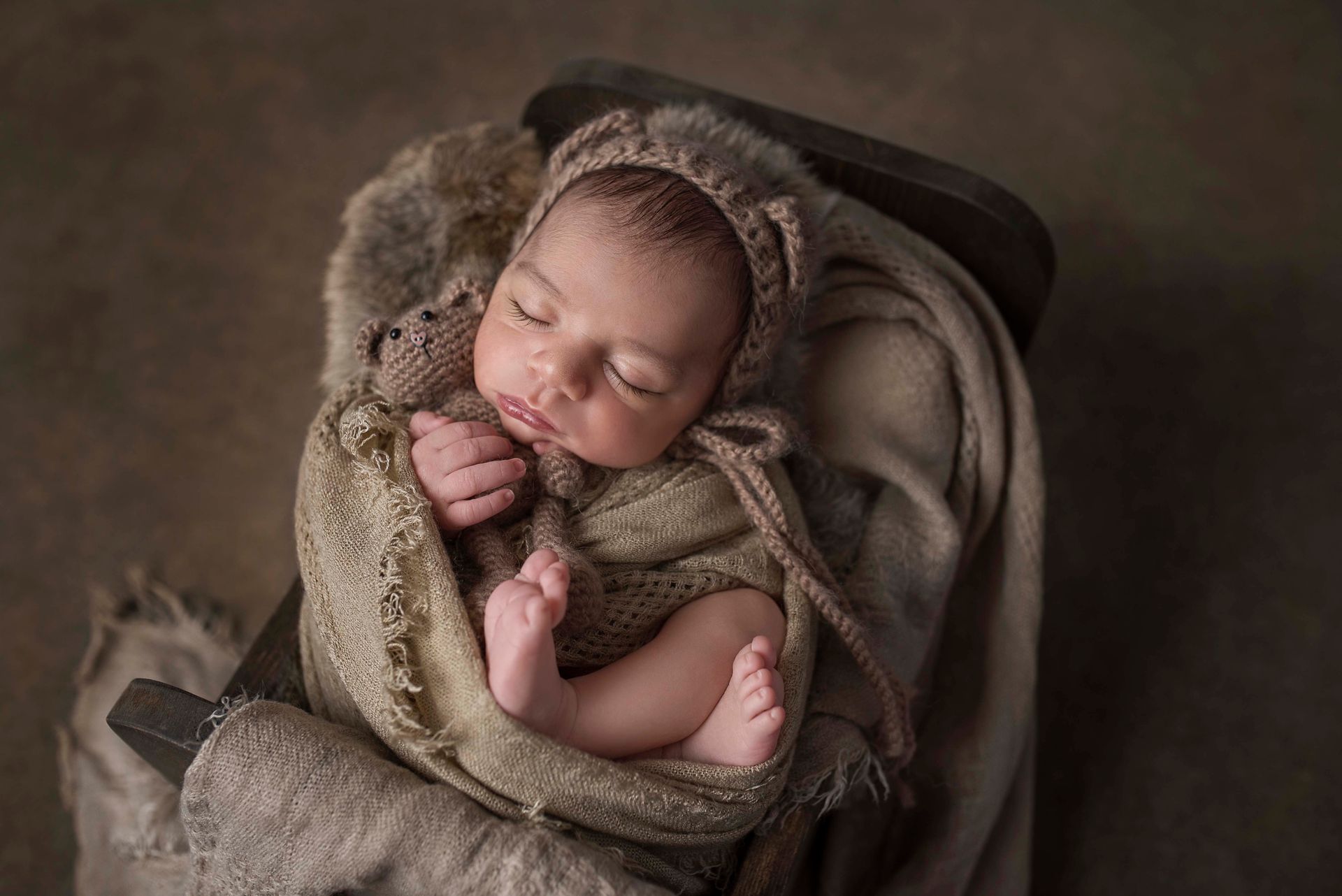 Newborn baby sleeping in a wooden chair, wrapped in a soft blanket and hat. Brown tones.