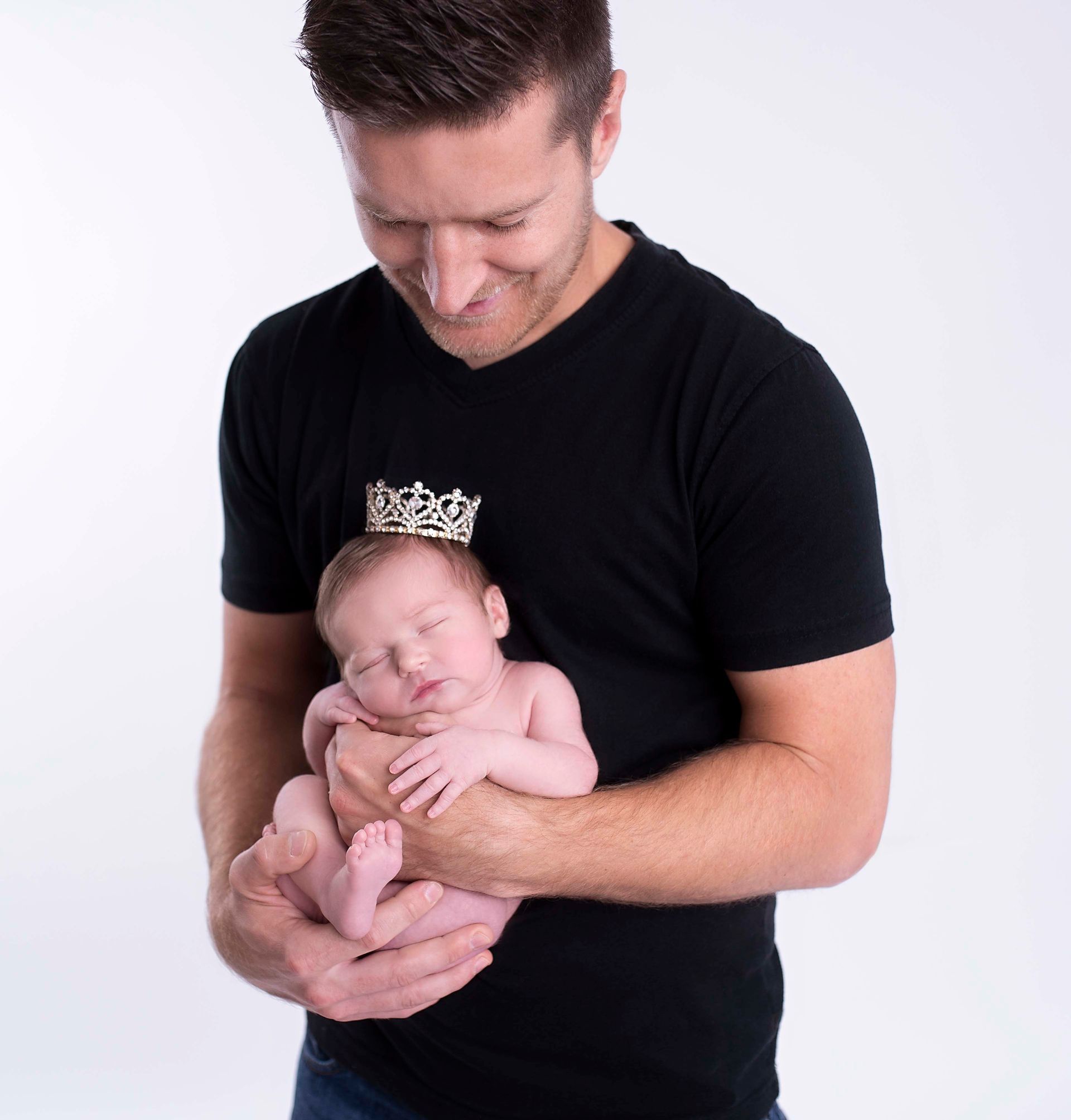 Man in black shirt holds newborn, looking down with a smile. White background.