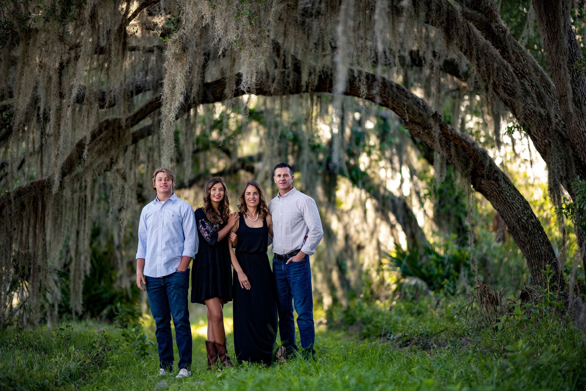 Family posing under a tree with Spanish moss; two women in black dresses, two men in button-up shirts and jeans.