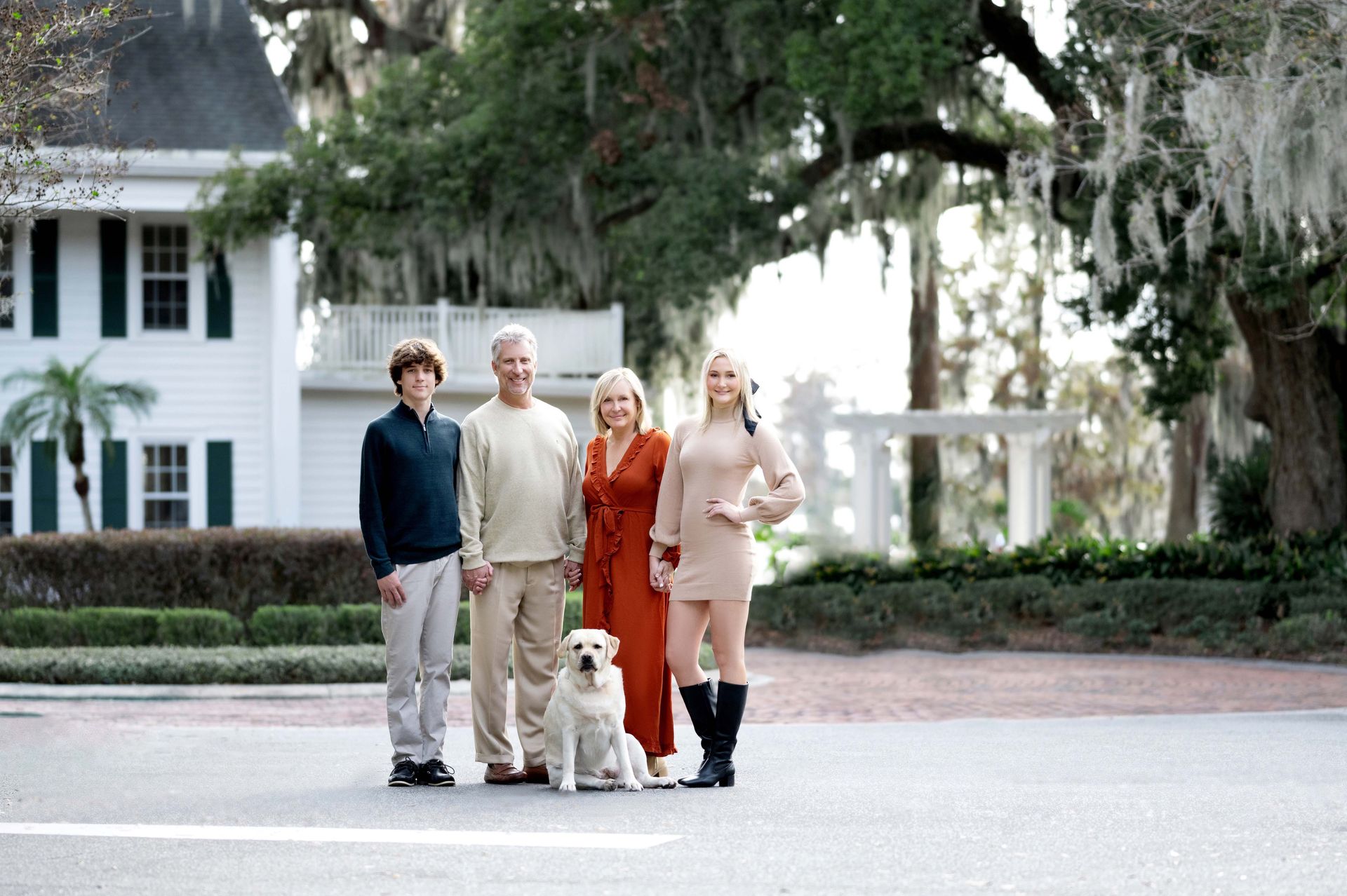 Family of four with a dog poses in front of a white house and a large tree, all looking at the camera.