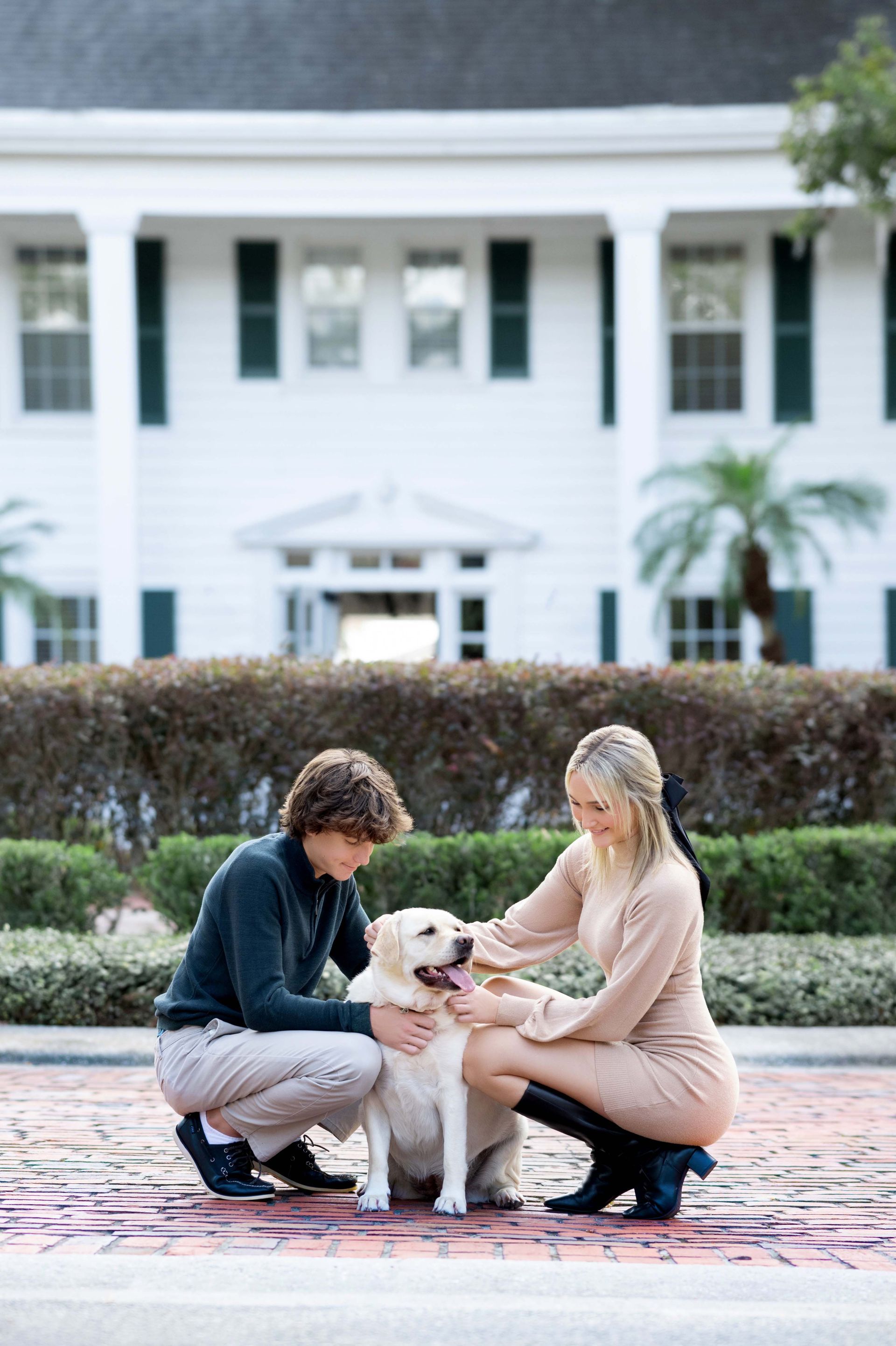 Couple petting a yellow lab on a brick path in front of a white building with columns.