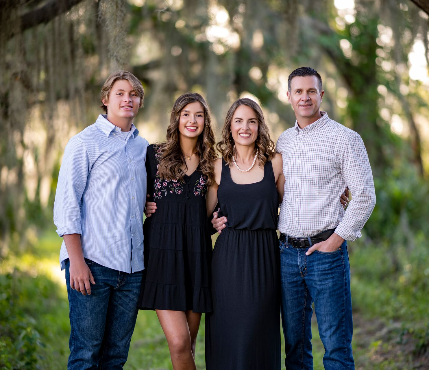 Family of four smiling, standing outdoors. A boy, girl, woman, and man. Green background.