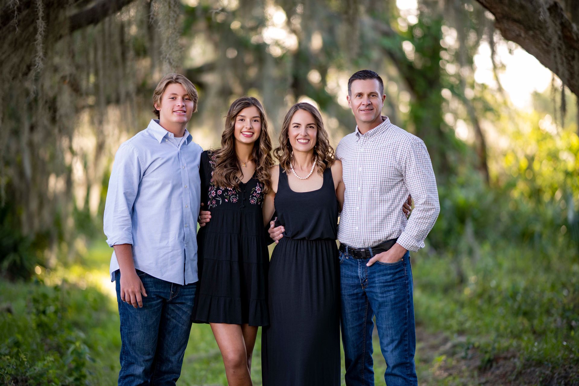 Family of four smiling, standing outdoors under a tree; two teens, parents, casual clothes.