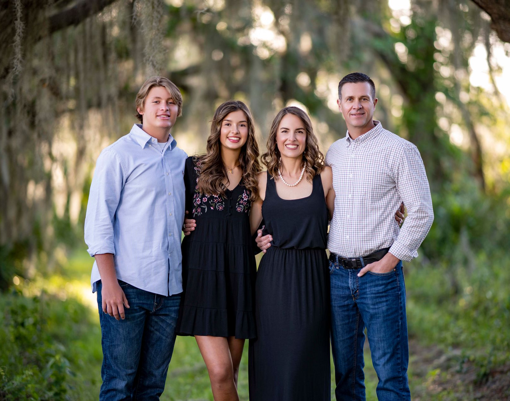 Family of four smiling outdoors: son, daughter, mom, dad, standing on a path with trees.